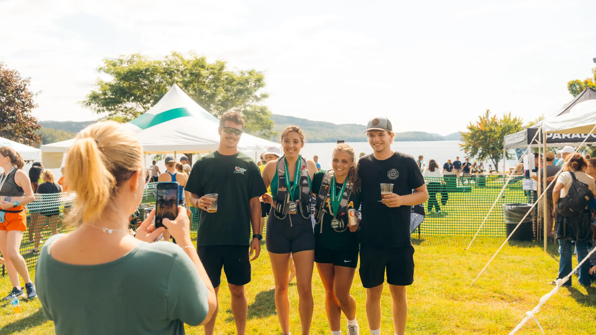 A woman takes a photo on her phone of four friends posing together with drinks and finisher medals.