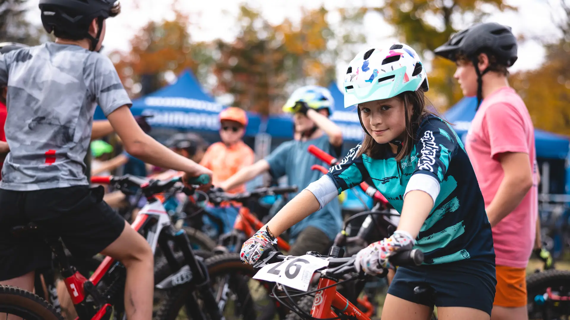 A young girl smiles while sitting on her mountain bike at a race, surrounded by other young racers.