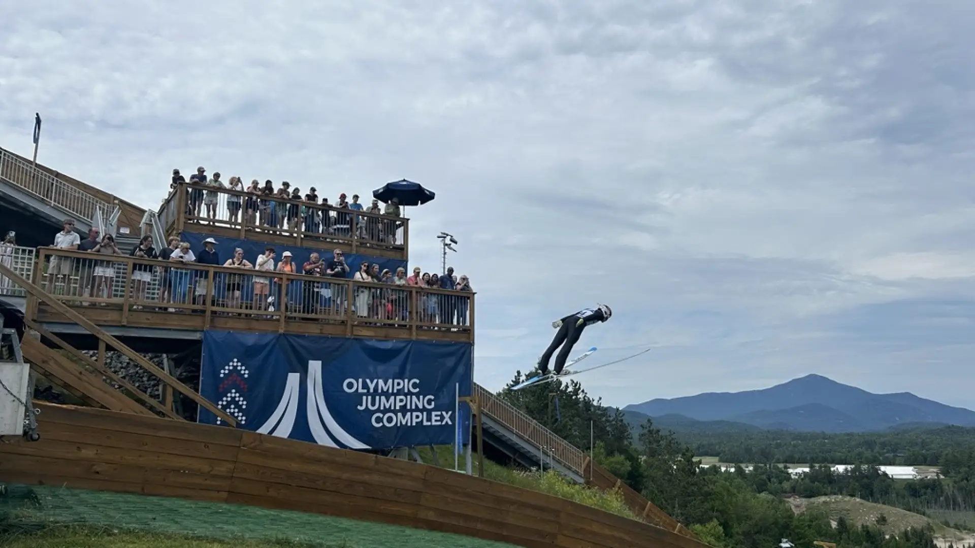 Ski jumper takes off from a lower jump over artificial turf while a two-level deck full of people looks on