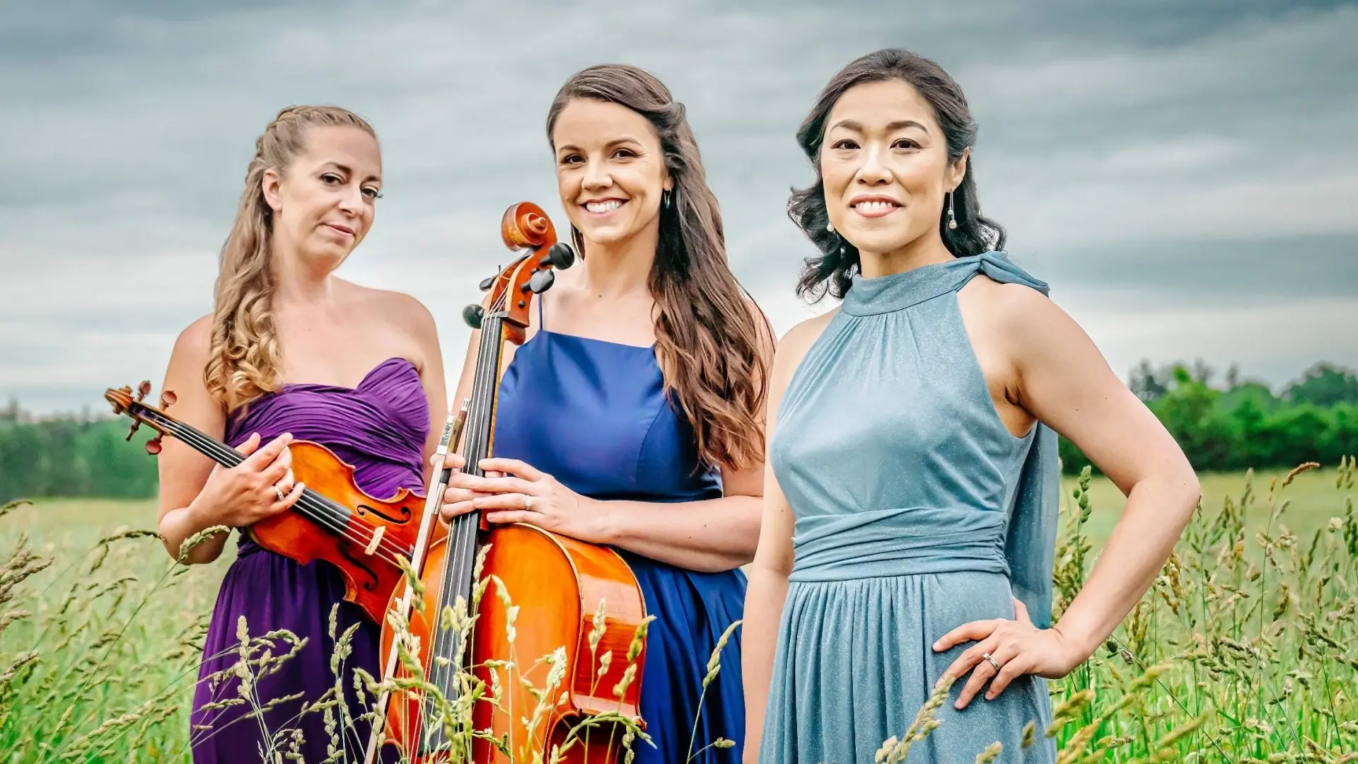 Three women in elegant dresses stand in a field. One holds a cello, another a violin.