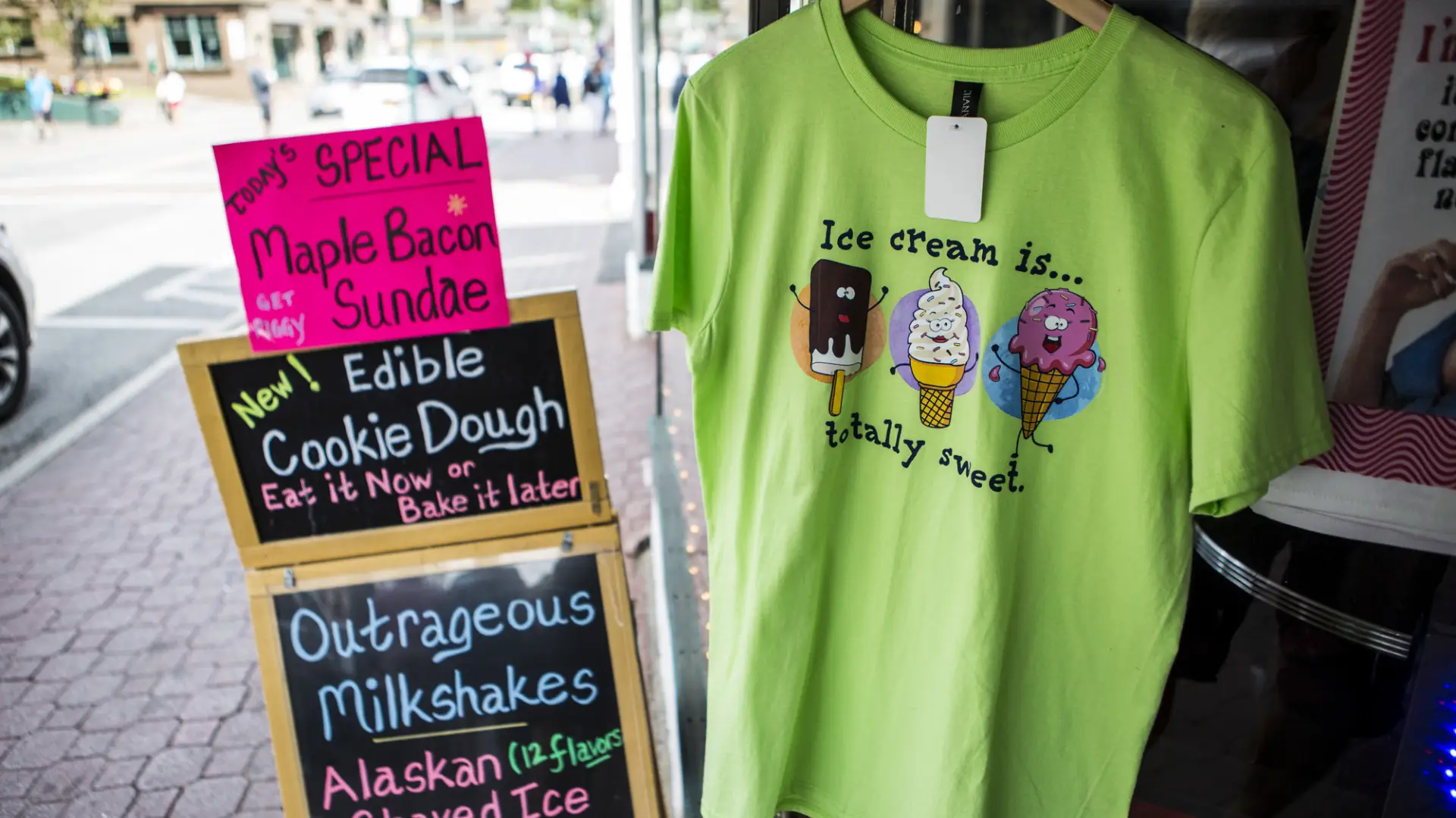 A sign advertising maple bacon ice cream sundaes sits next to a tshirt outside a shop.