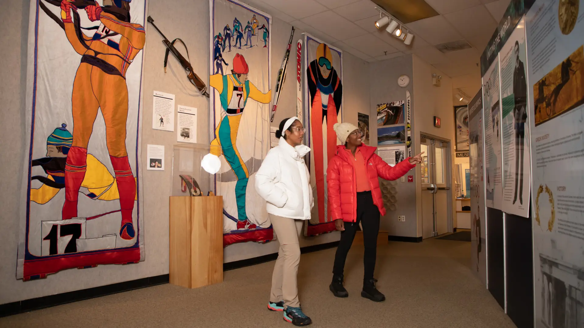 Two women in white and red jackets laugh and walk through an Olympic history museum. .