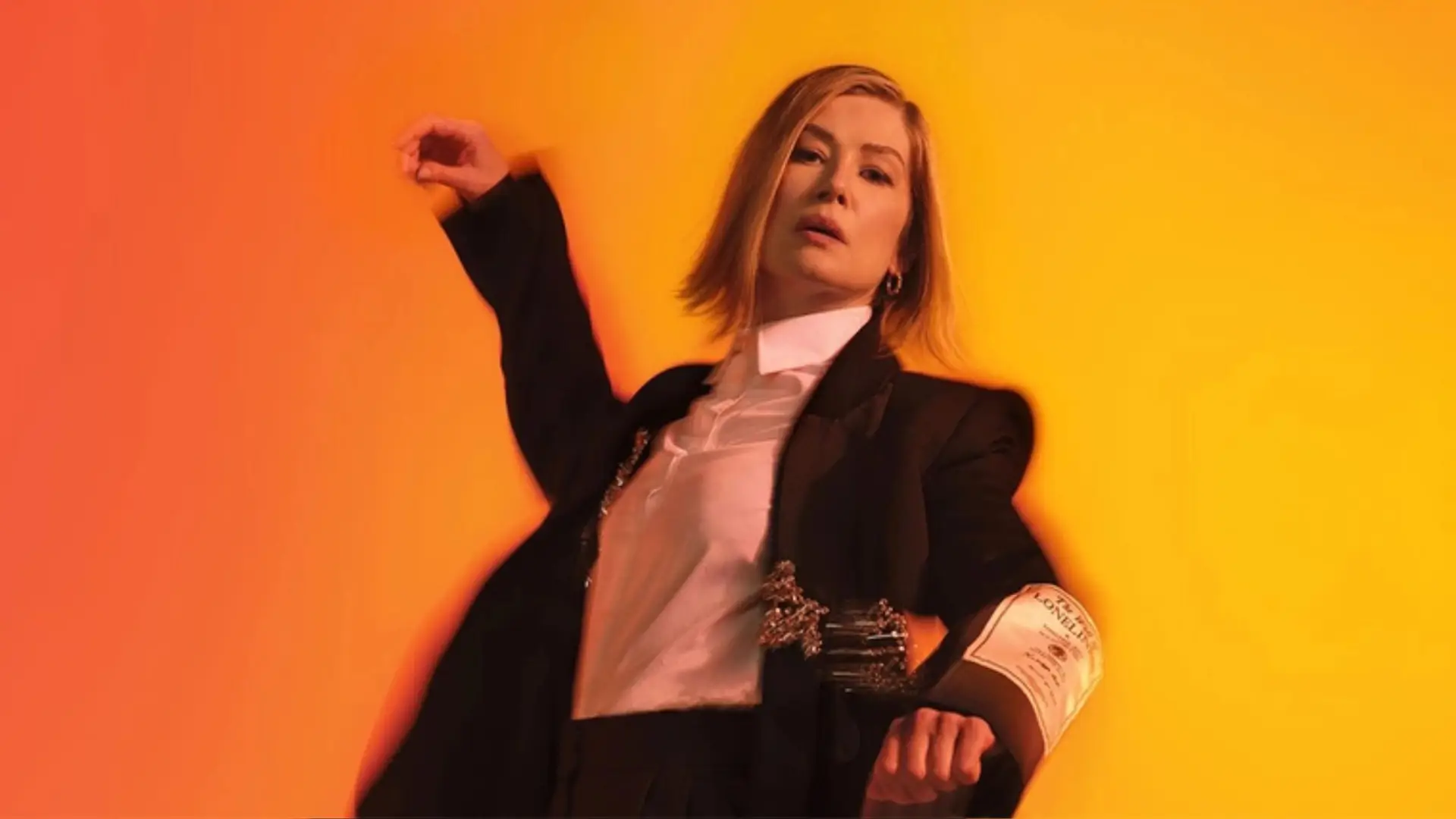 Rosemund Pike in a black and white suit dancing in front of yellow/orange background