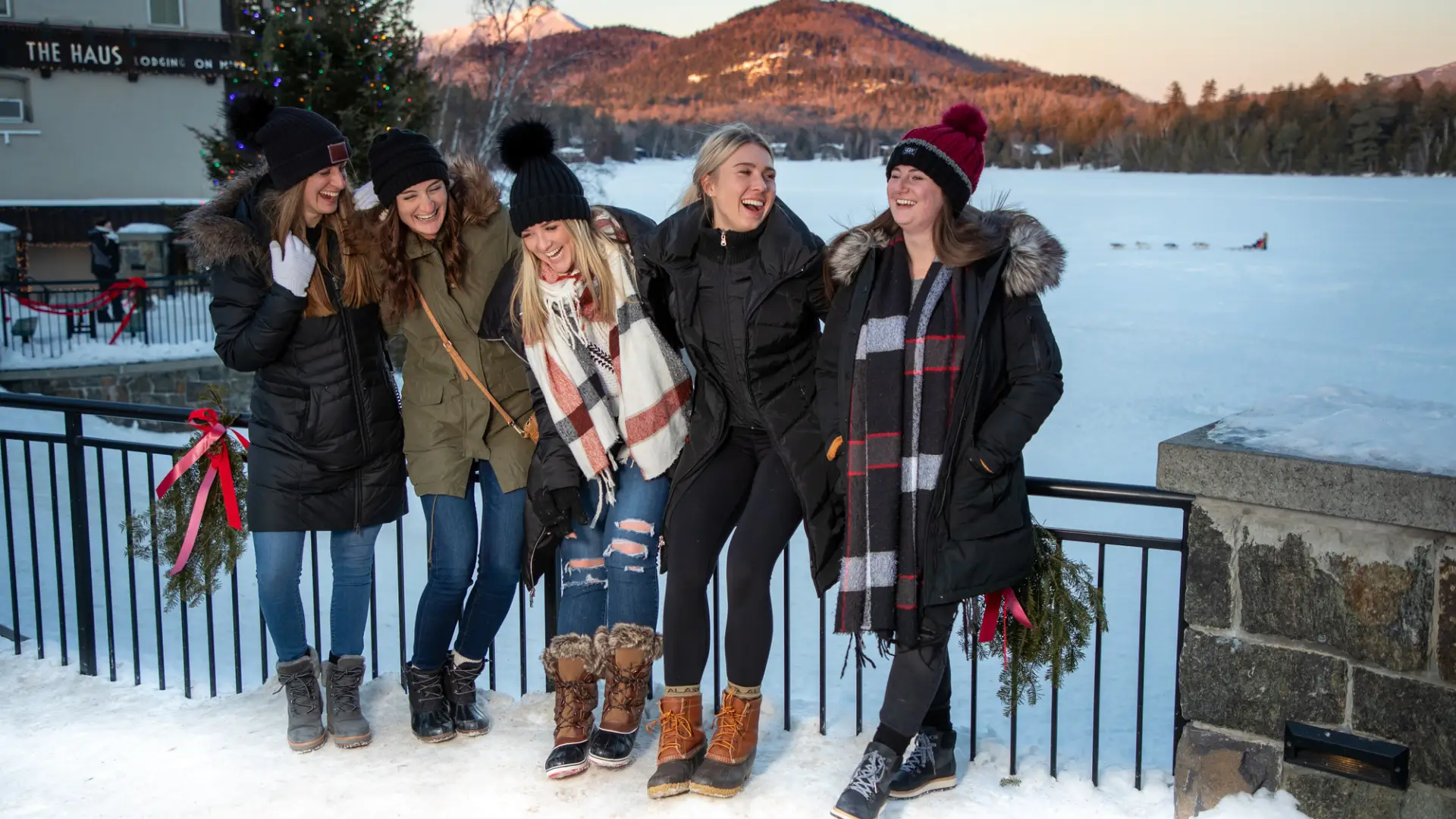 Five women in winter clothing laugh in front of a snowy lake and sunset-lit mountains beyond.