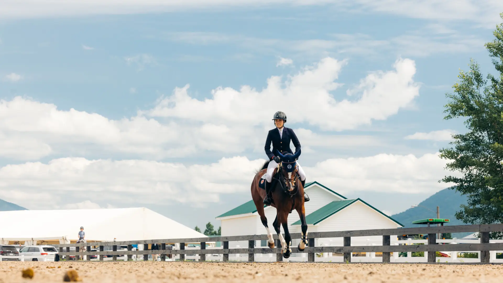 Equestrian competitor rides horse on dirt ring with white and green roofed building in background