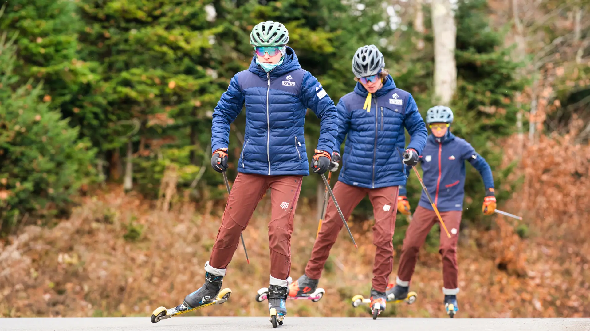 Three teenage student athletes practice cross-country skiing on specially equipped skate skis on a forested road.