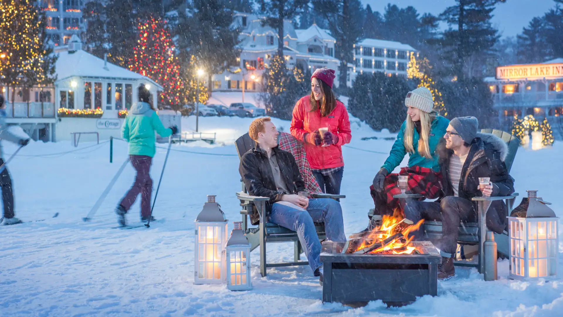 Happy people sit and chat around a fire pit with the glow of christmas lights dot the background and a skiier slides by.