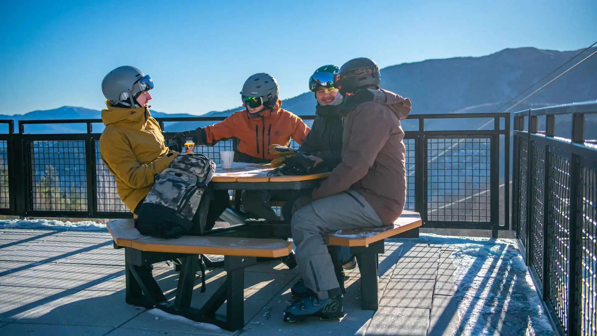 Four friends smile and laugh at an outdoor picnic table on a patio while dressed in ski clothes.