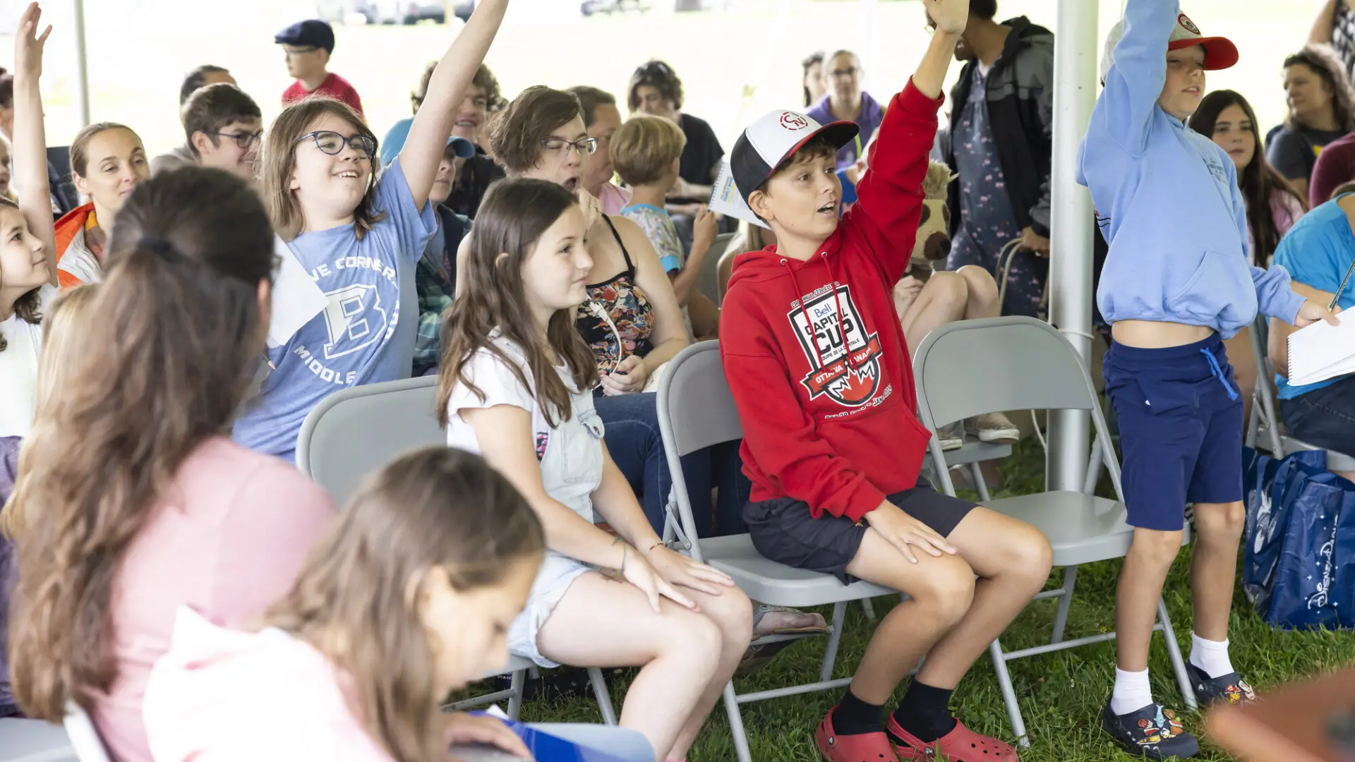 Collection of kids sit in folding chairs under outdoor tent at a presentation, some raising their hands