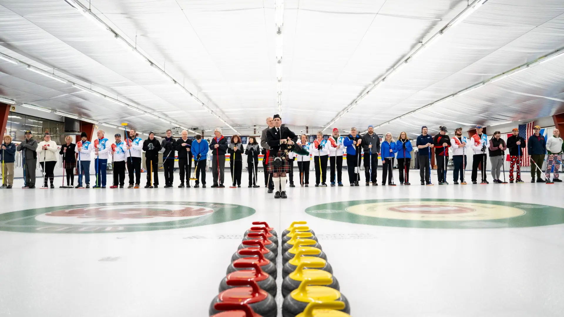 A piper in traditional attire plays bagpipes, surrounded by a crowd of curlers at a curling event during Empire State Winter Games, with colorful stones lined up in the foreground.
