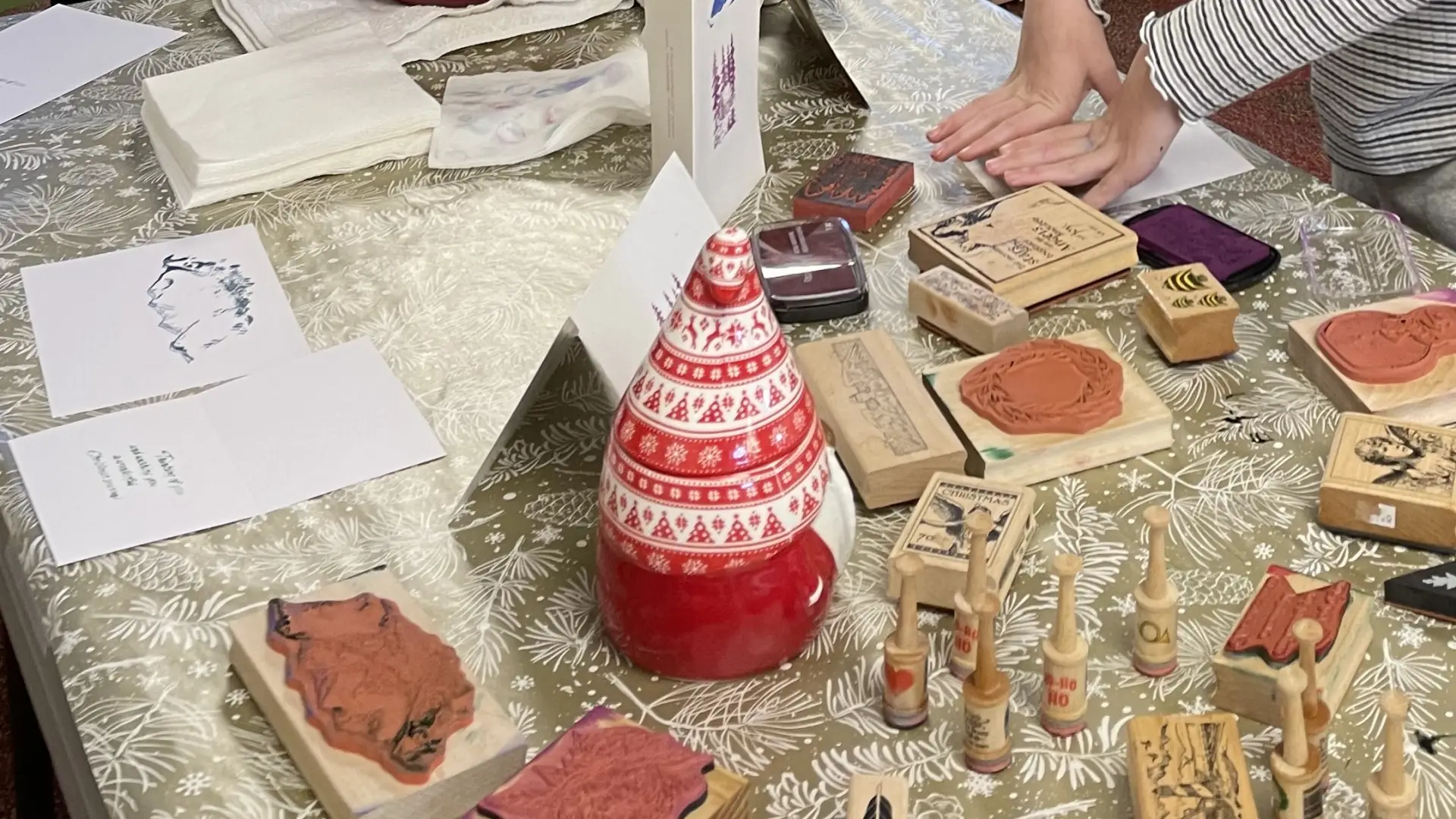 Table covered in various stamps, paper, a Santa-shaped ceramic decoration, and small hands pressing a stamp down on paper