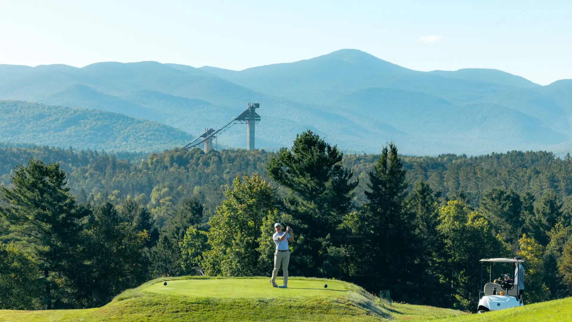 A man hits a golf ball in front of Olympic ski jumps. 