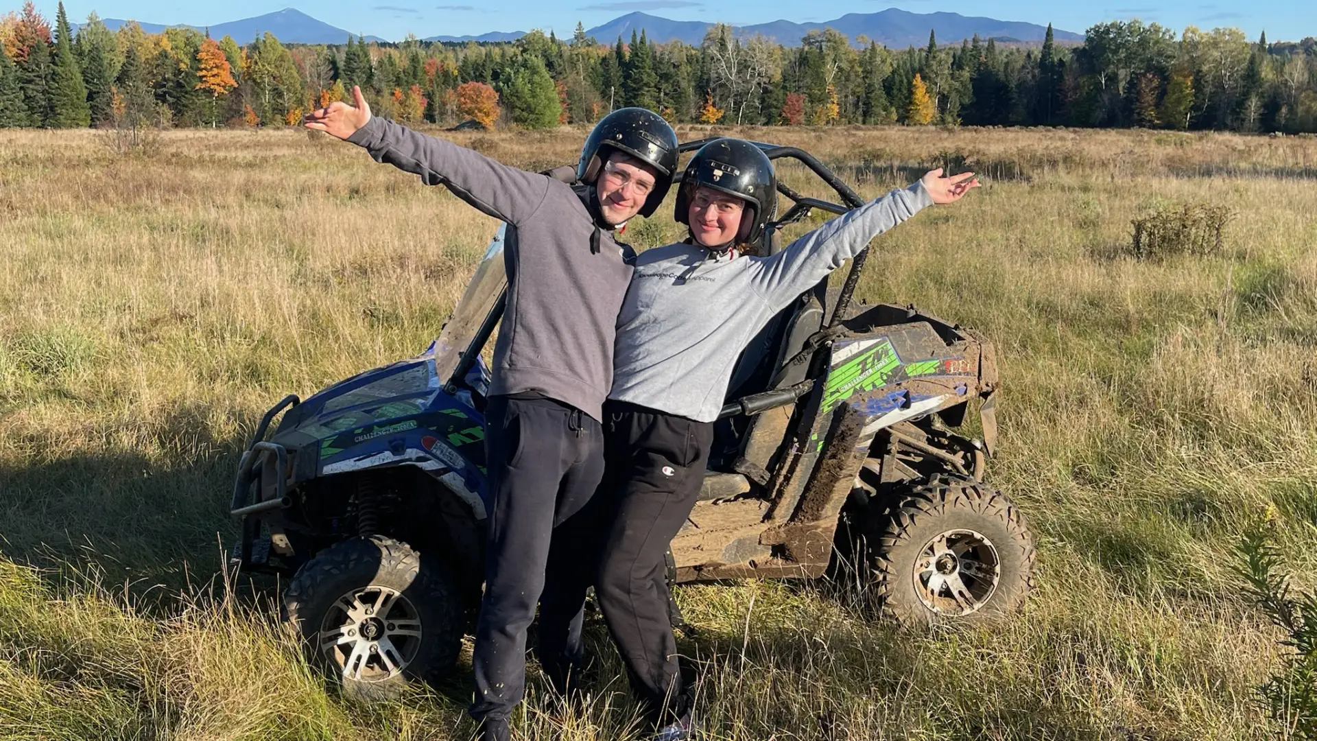 A happy couple smile into the camera with an arm raised in excitement in front of a UTV parked in a grassy meadow with tall pine trees and the Adirondack Mountains in the background under a wide blue sky.