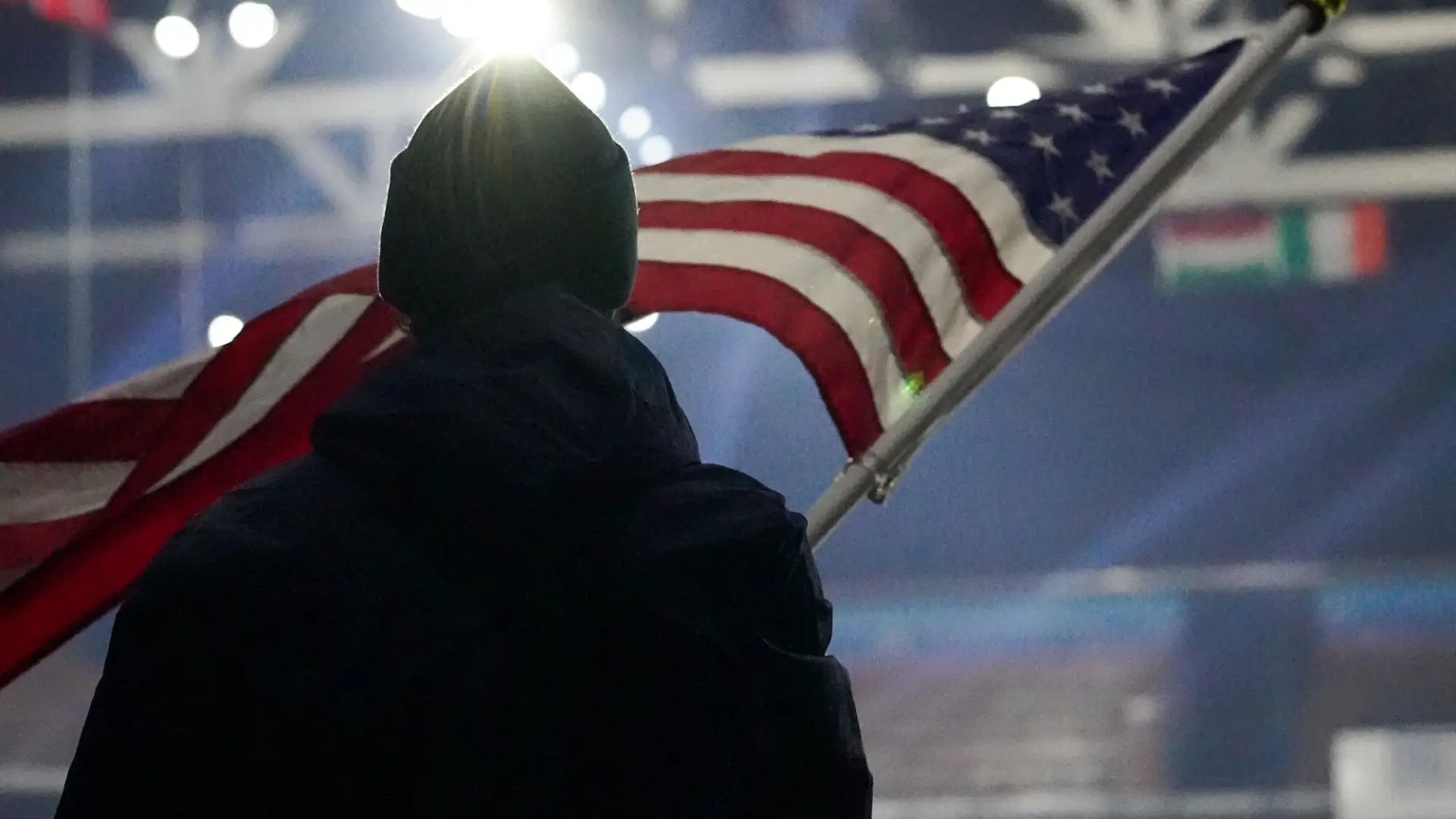 Person stands facing away from camera in a well-lit ice rink holding an American Flag on a pole