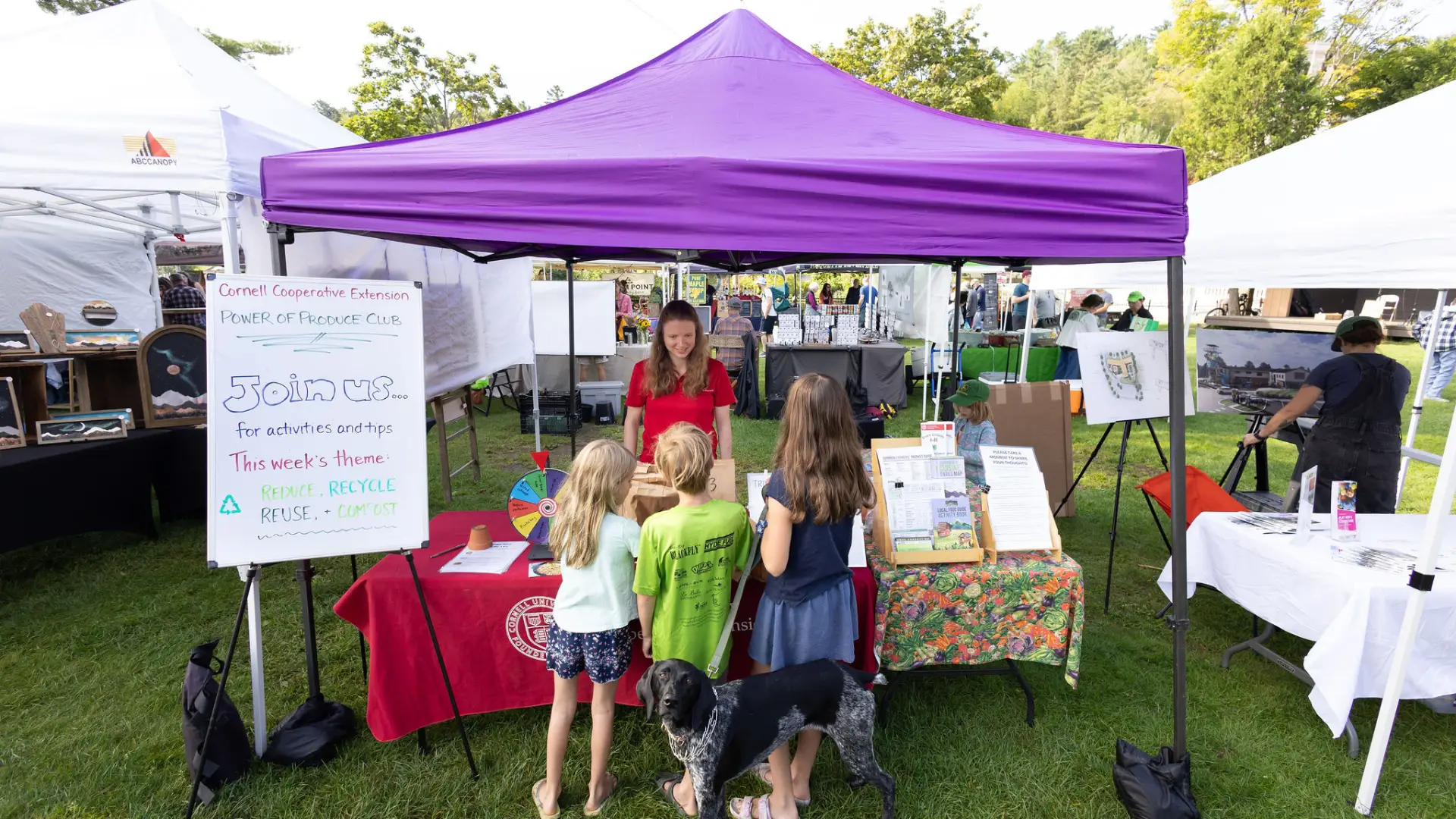A purple tent at an outdoor event with a table displaying information about the Cornell Cooperative Extension's Power of Produce Club.