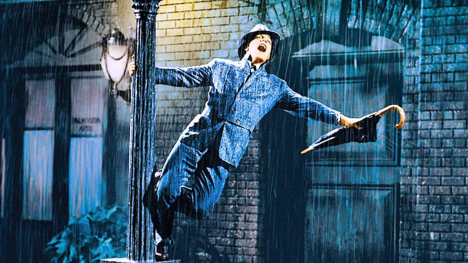 A man in a gray suit and hat, Gene Kelly, joyfully swings from a black lamppost during a heavy rainstorm.