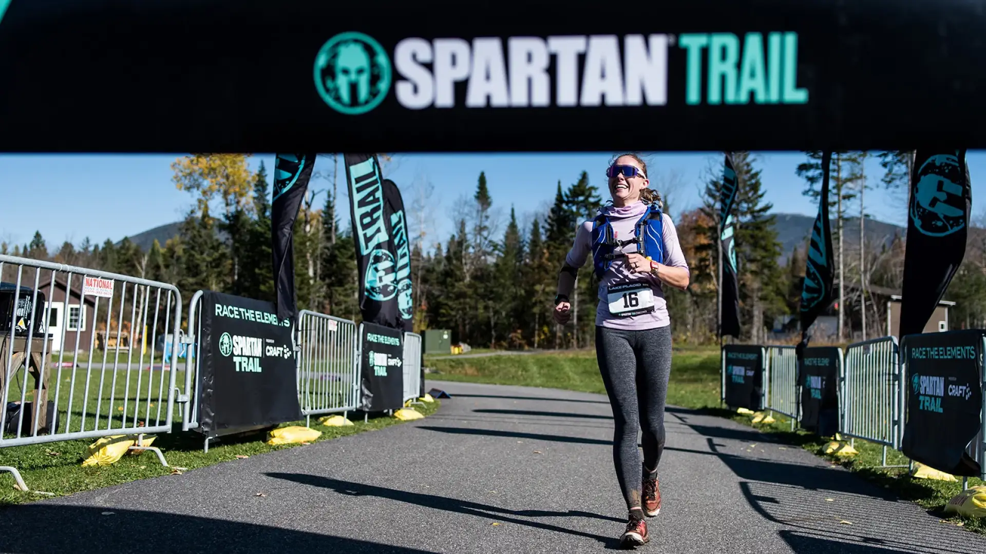 Woman wearing running vest smiles as she runs beneath a Spartan Trail arch over a paved path past event fencing