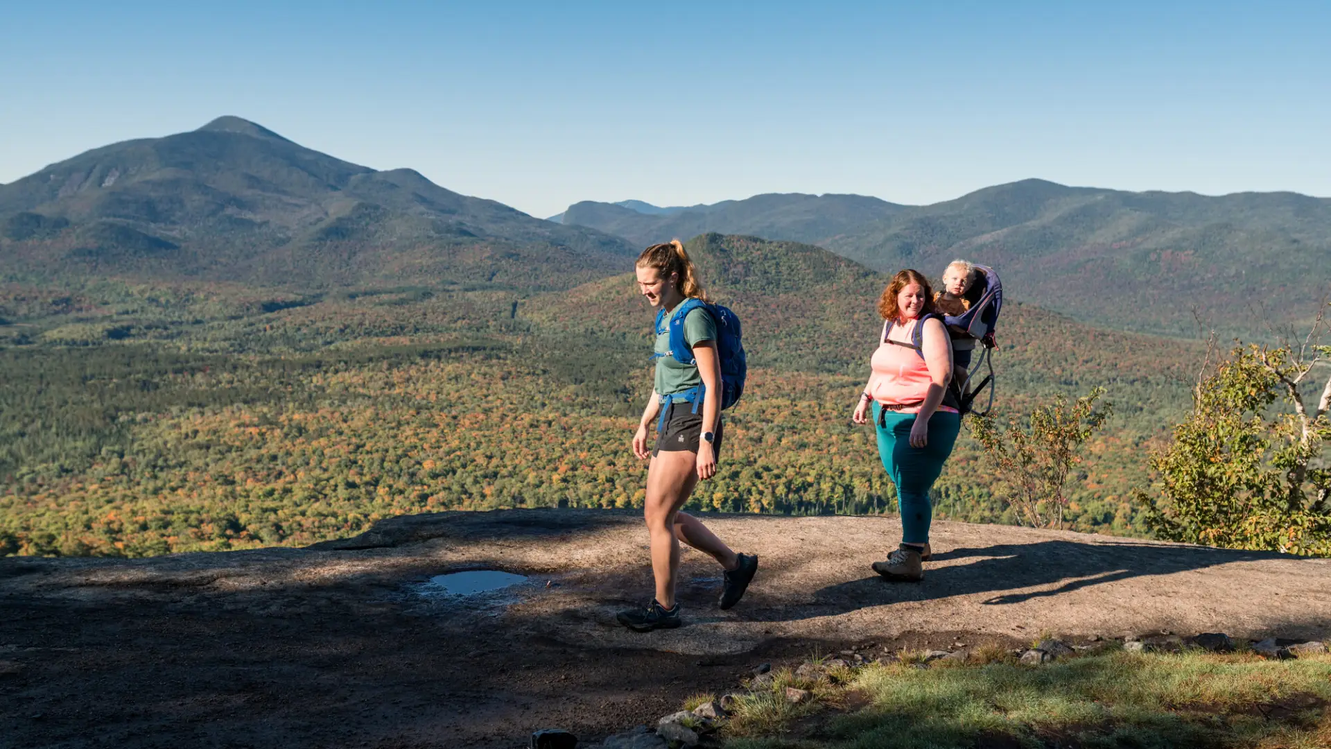 Two women and a baby at the summit of Mt. Van Hoevenberg in early fall overlooking the mountain range.