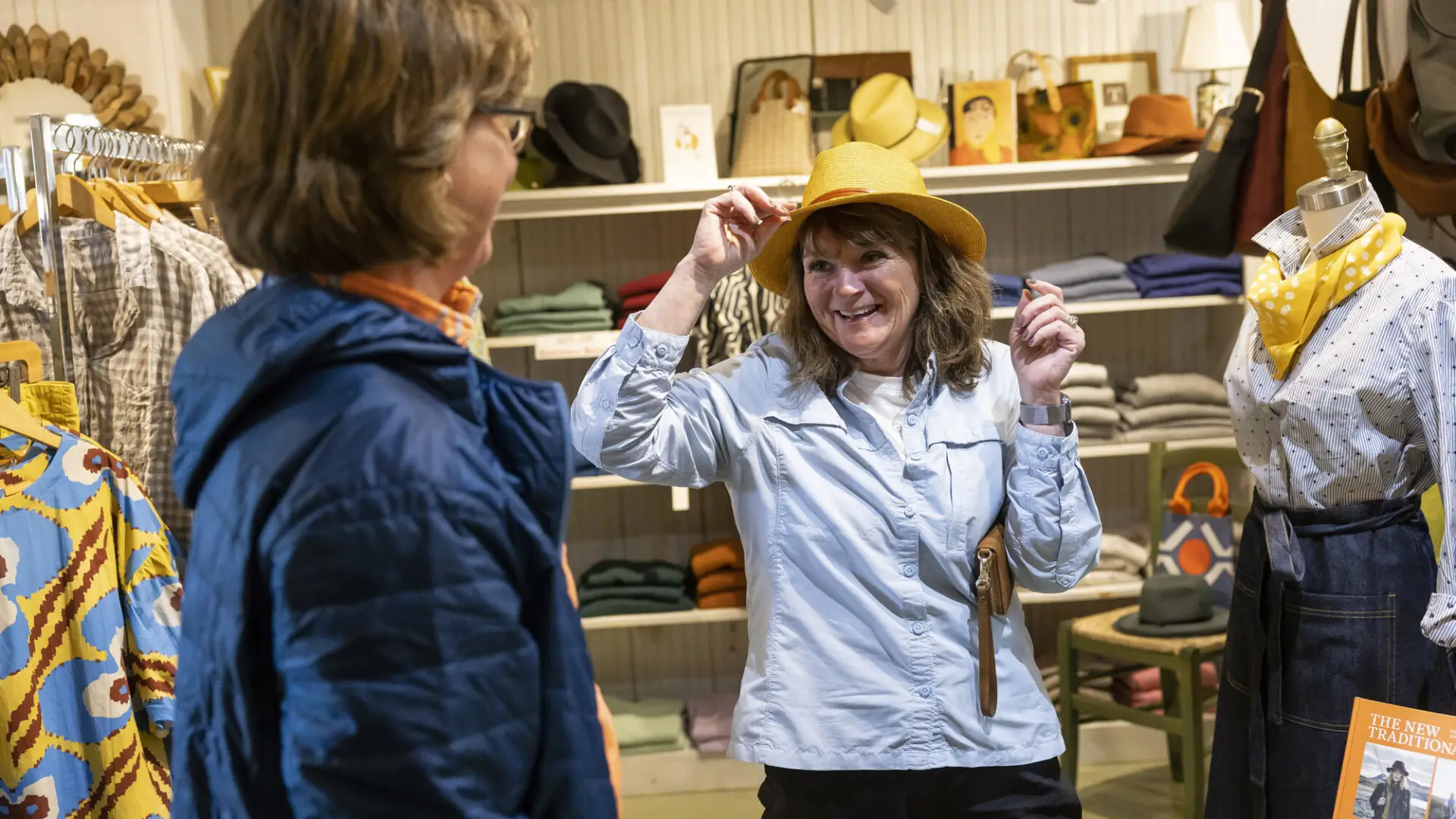 A jovial woman tries on a fun, yellow hat infront of a beautifully curated outfit in the women's section of The Birch Store
