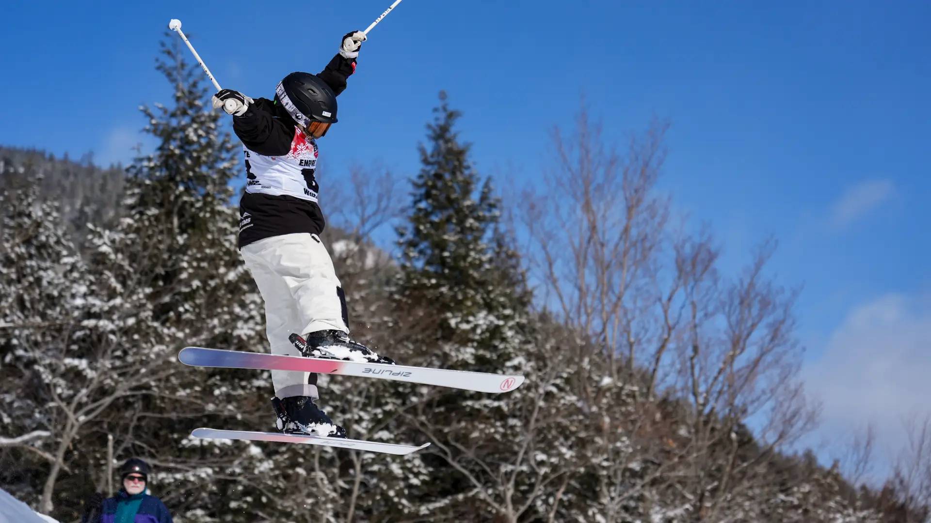A freestyle mogul skier performs a jump with poles extended against a clear blue sky and snow-dusted evergreens.