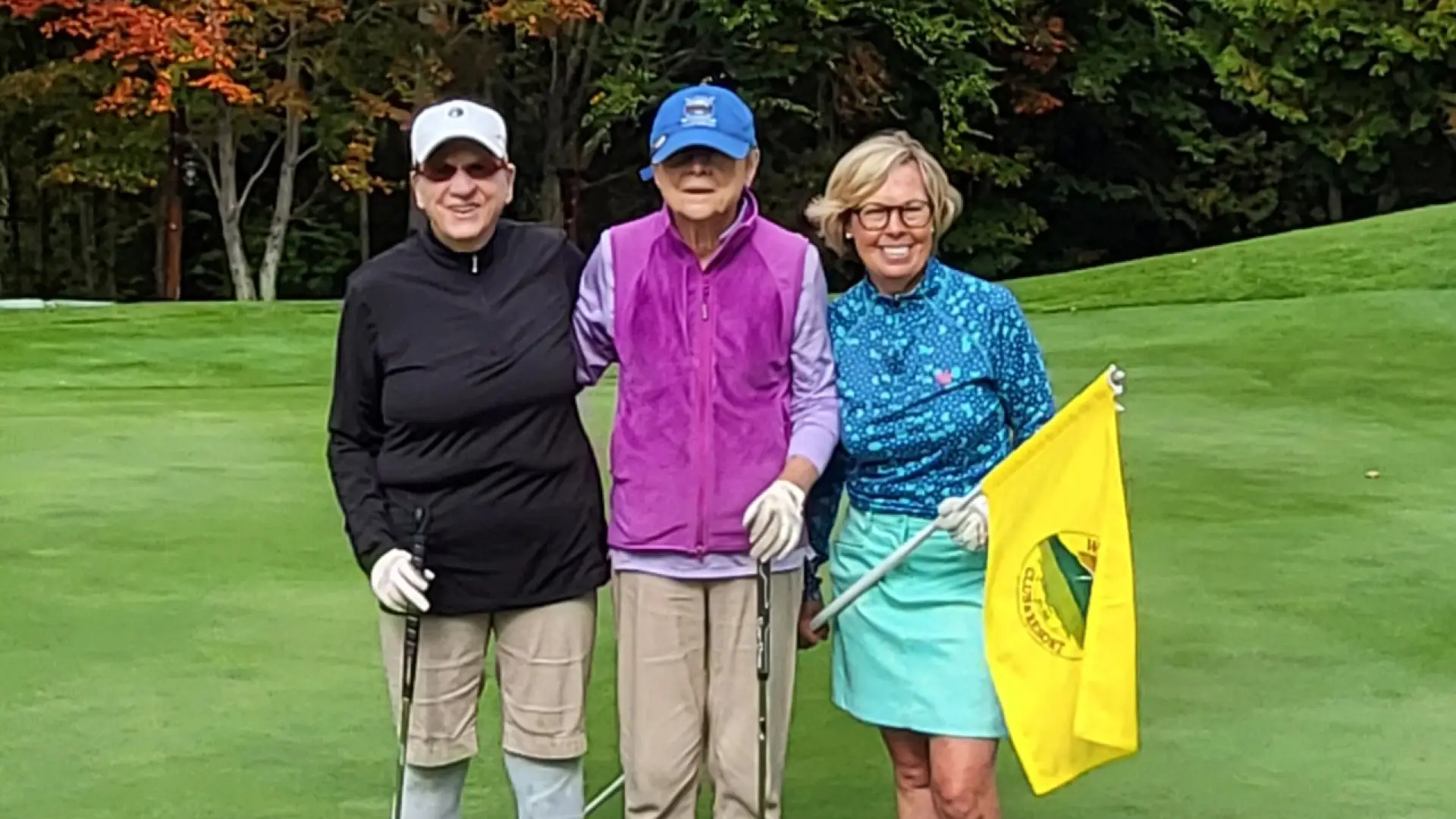 Trio of golfers stand together posing for camera on golf course, two holding golf clubs and one holding a yellow flag