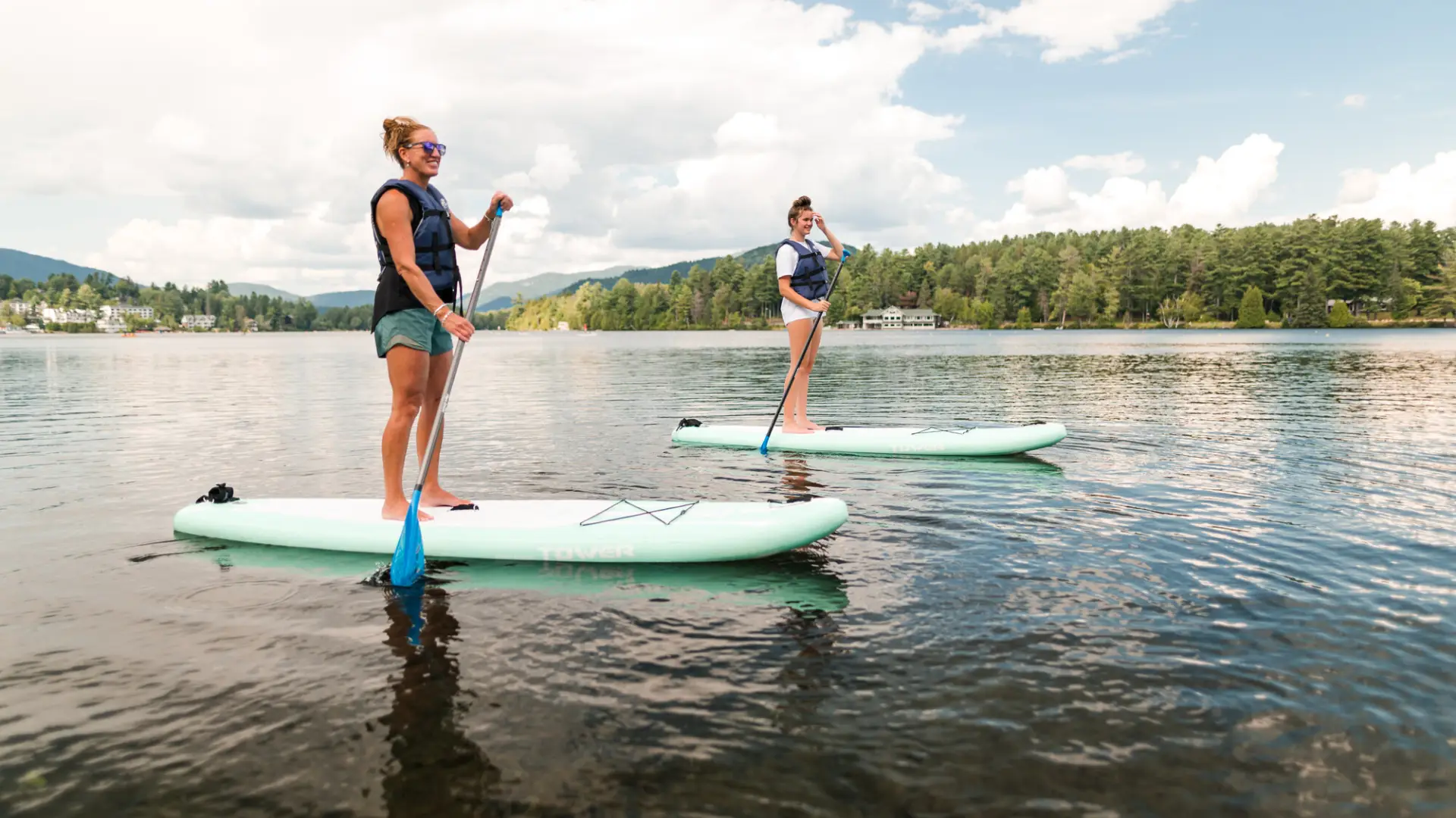Two women paddle standup paddleboards on a calm lake on a sunny day. Low hills and trees line the shoreline in the background.