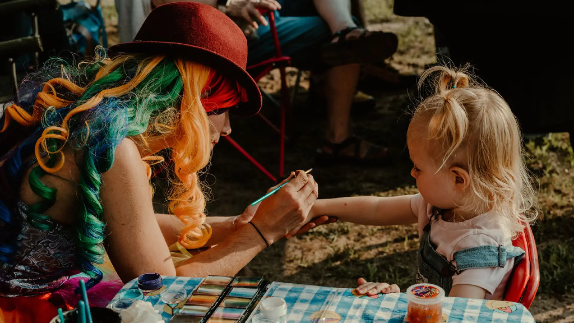 Woman wearing rainbow colored wig paints on the hand of a small child with art supplies on table in front of them