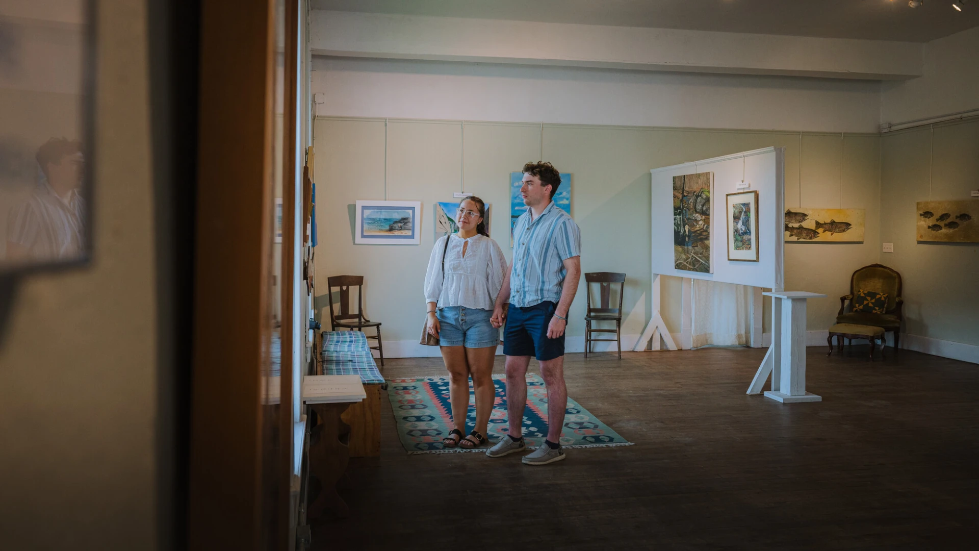 Man and woman stand holding hands in open concept art gallery in natural light looking at art wearing summer clothes