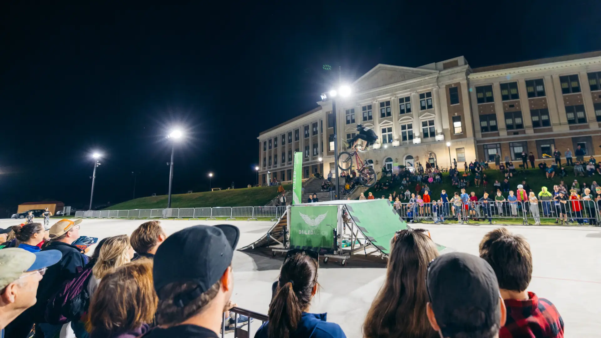 Stunt rider flips in mid-air over temporary jump on the track of the Olympic Oval at night as crowd looks on