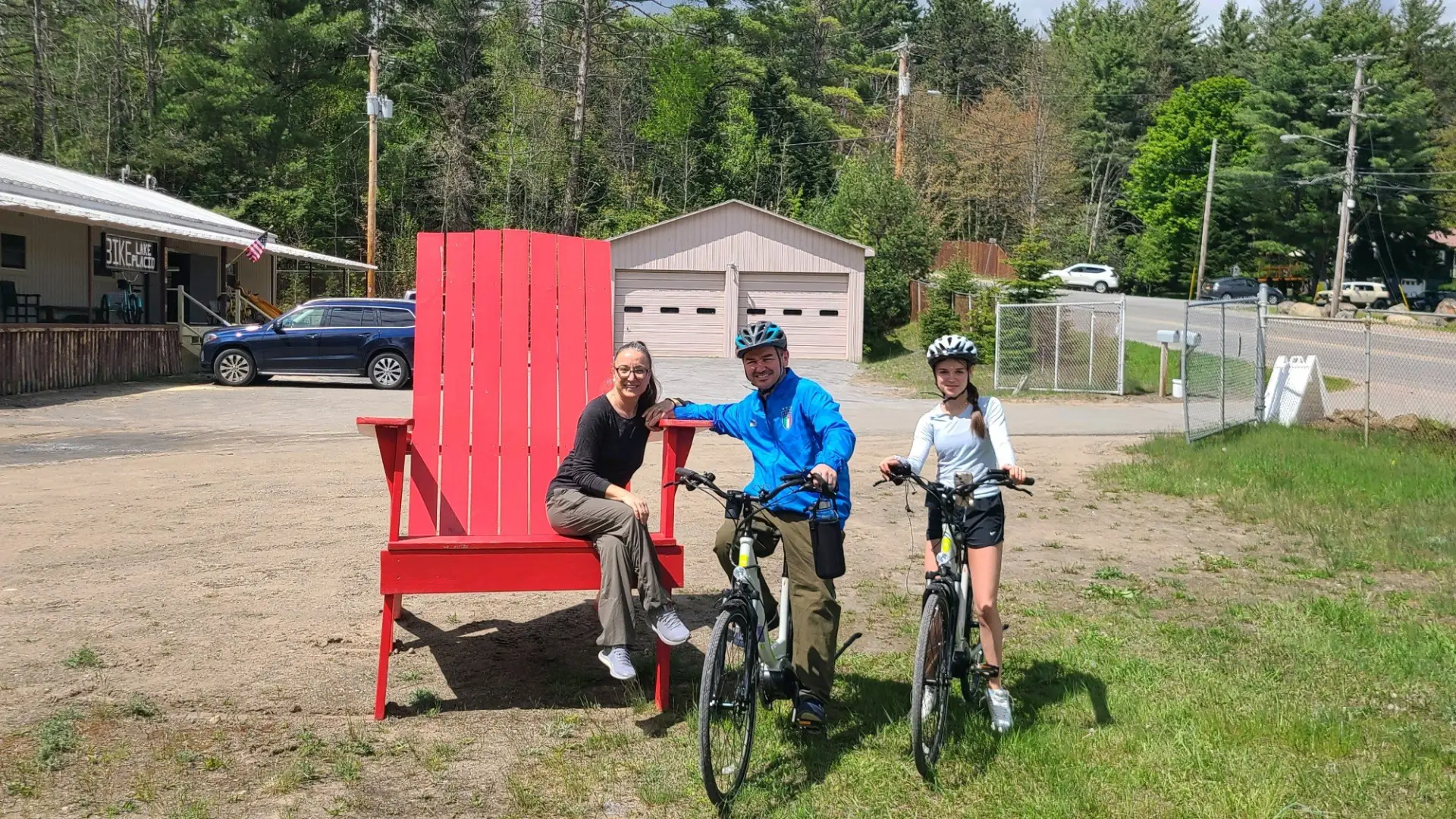Three smiling people surround the giant red adirondack style chair out front of Bike Lake Placid