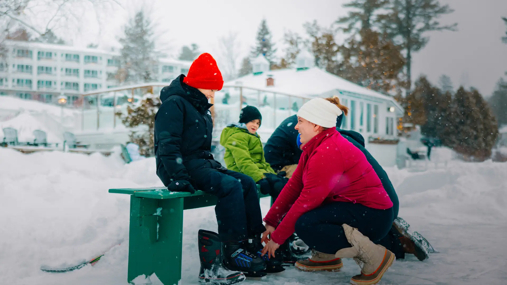 Woman helps child sitting on green bench put on ice skates on frozen lake