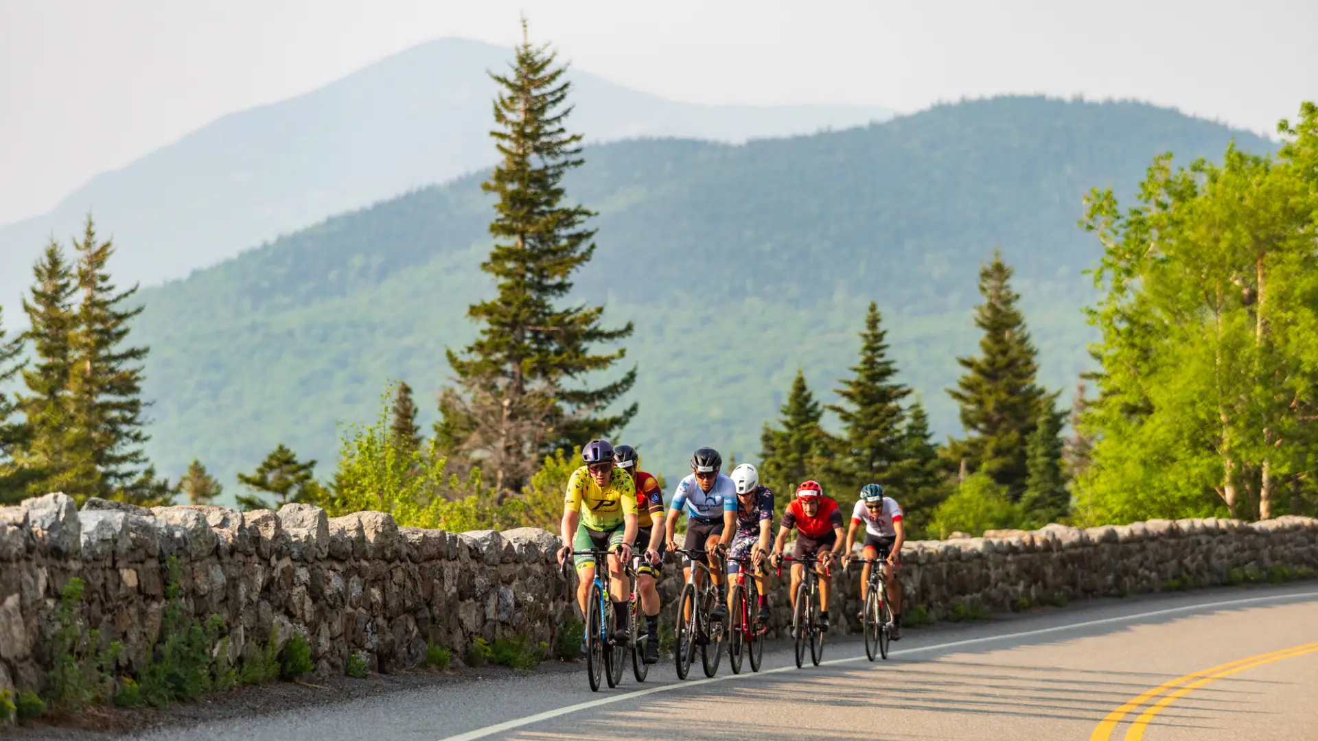 A group of cyclists rides in a tight pack along a winding mountain road bordered by a stone wall.