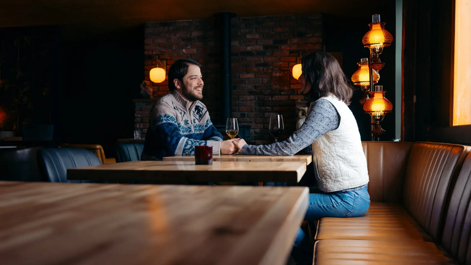 Couple sits at a wooden table in a restaurant with a dark, moody ambiance drinking wine