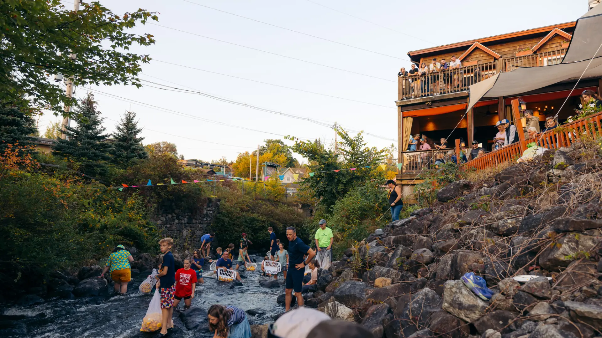 Children pick yellow rubber ducks out of a river while people look on from a restaurant deck up the embankment 