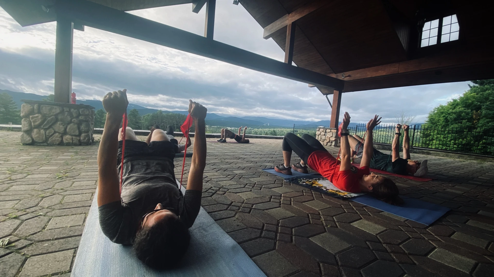 People lay on backs on yoga mats pushing arms up with resistance bands in workout class held outdoors on covered brick patio