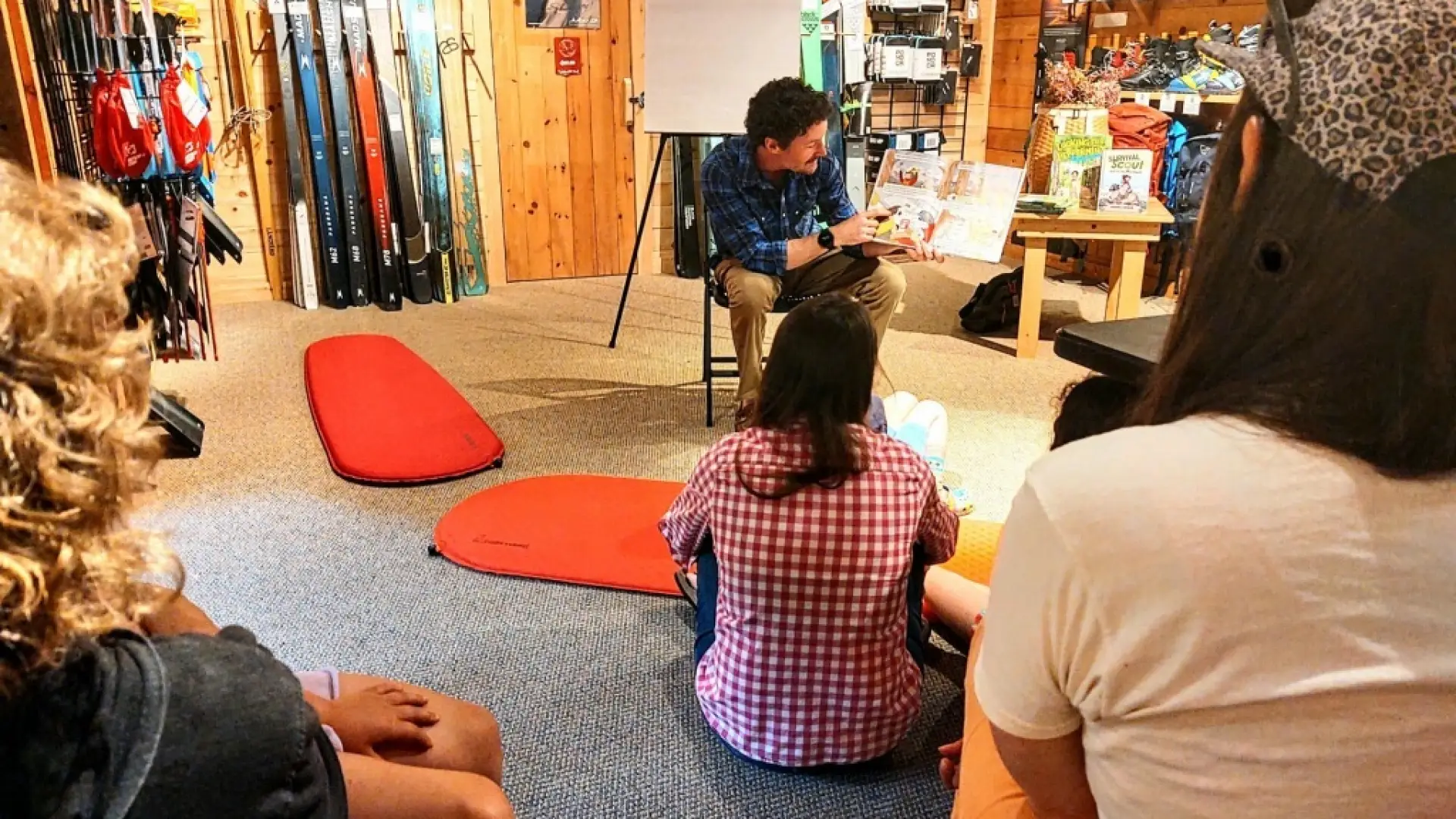 A man reads a children's book to a group of children sitting on the floor. Ski equipment is visible in the background.