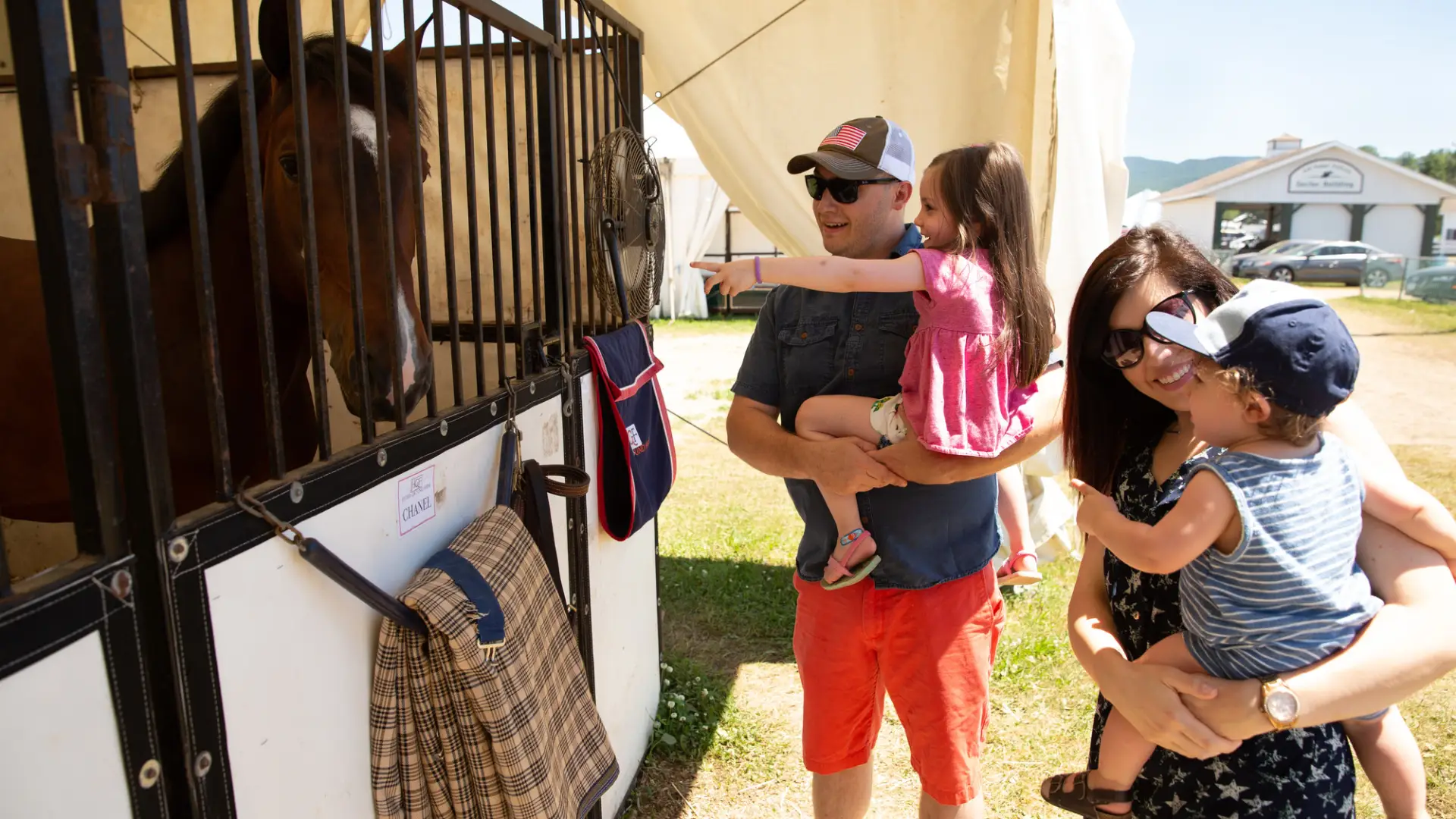 Father holds young daughter and mother holds baby boy as they look at horse through stall grate at Lake Placid Horse Show