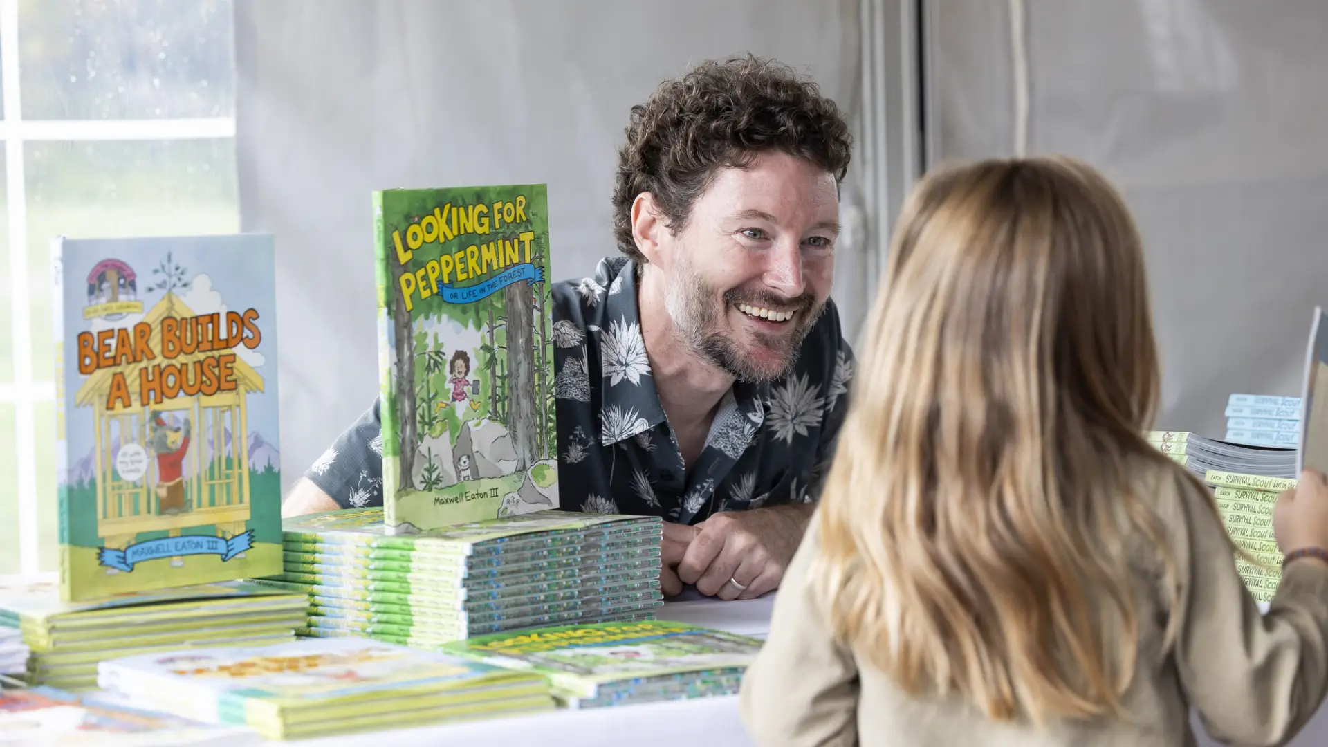 Middle aged white male author sits behind book display on table smiling at child standing across from him
