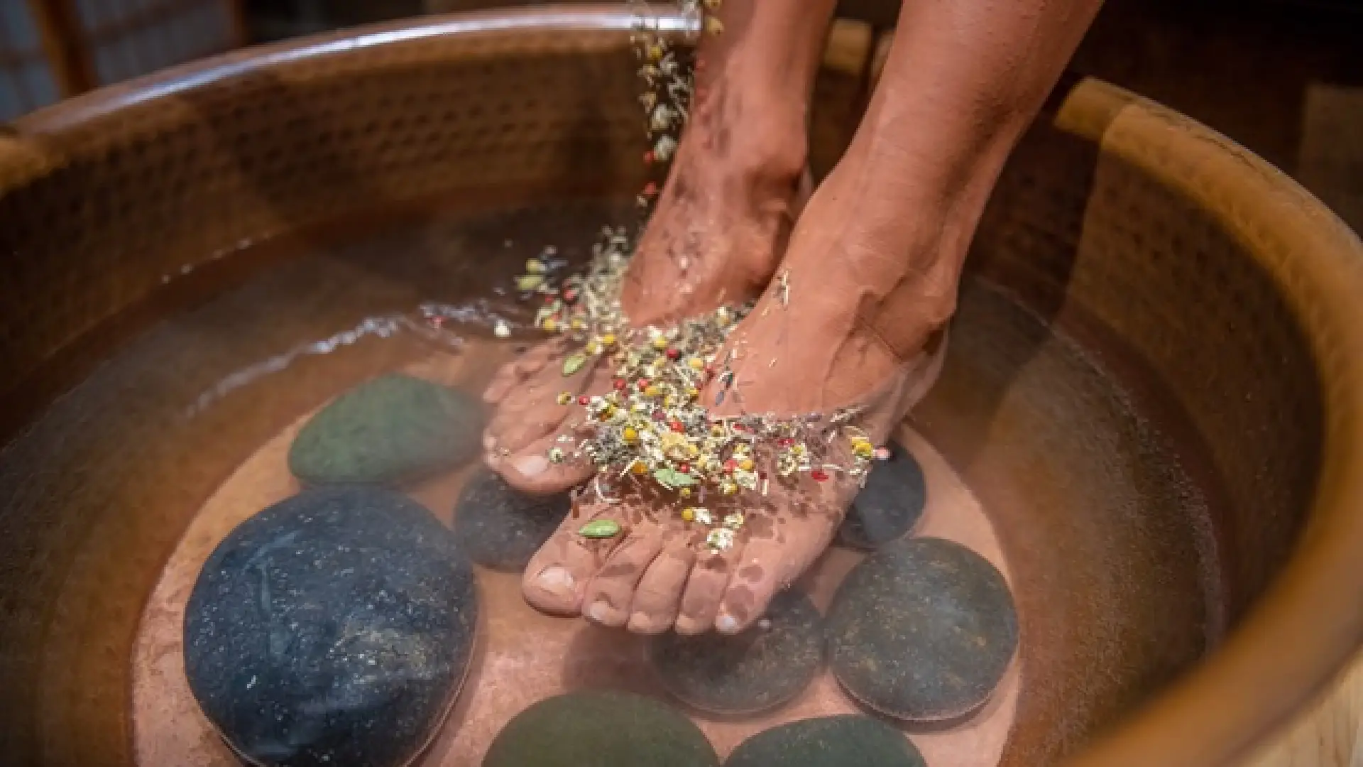 Feet immersed in a tub of steaming water, topped with herbs and some river stones within