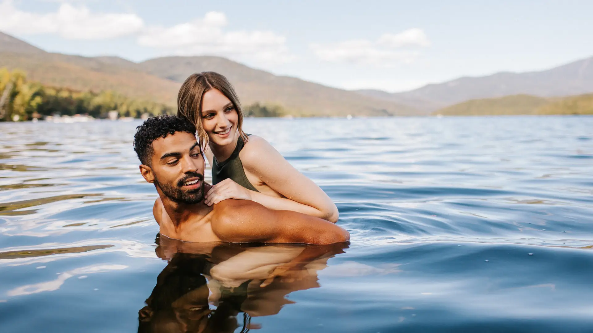 Man holds woman on his back in lake water with mountain background