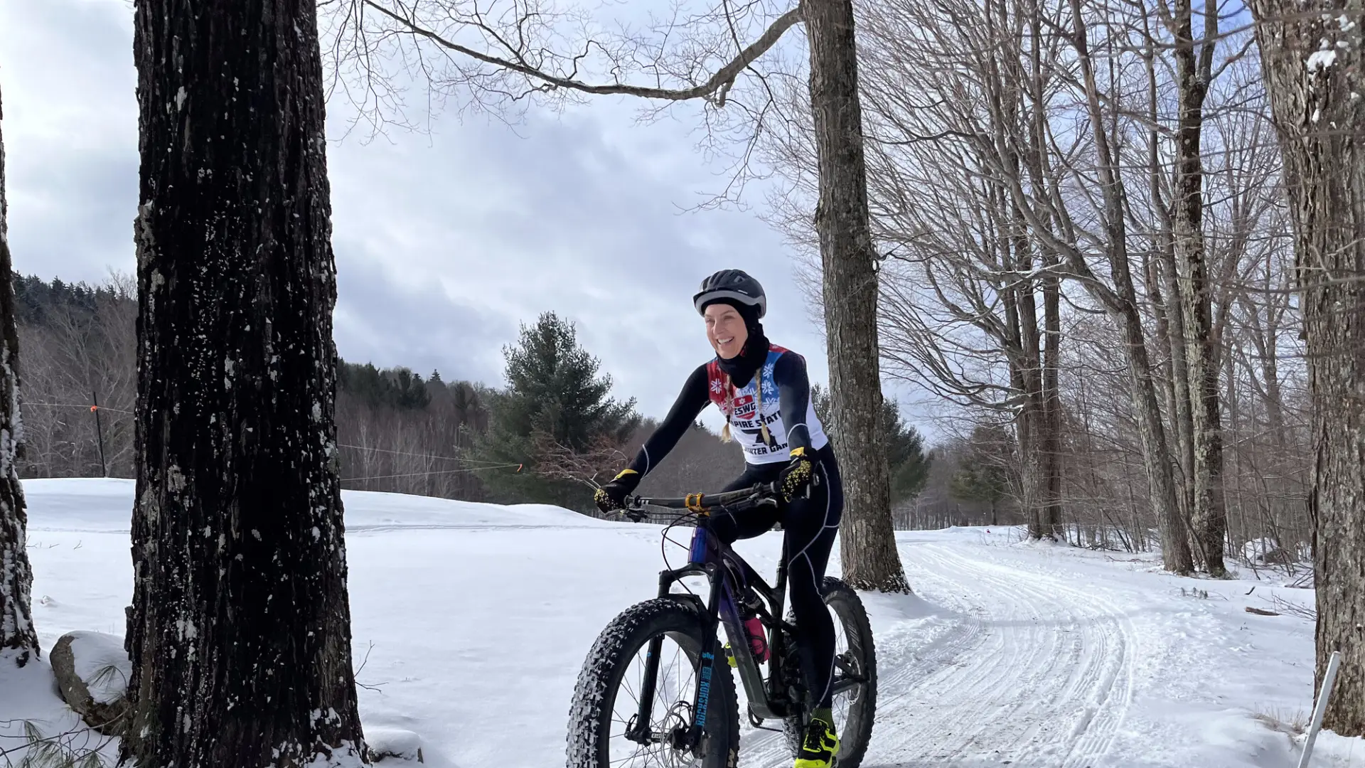 Person biking with a big smile on a snowy trail through a forest, wearing winter gear and helmet. The scene conveys excitement and adventure.