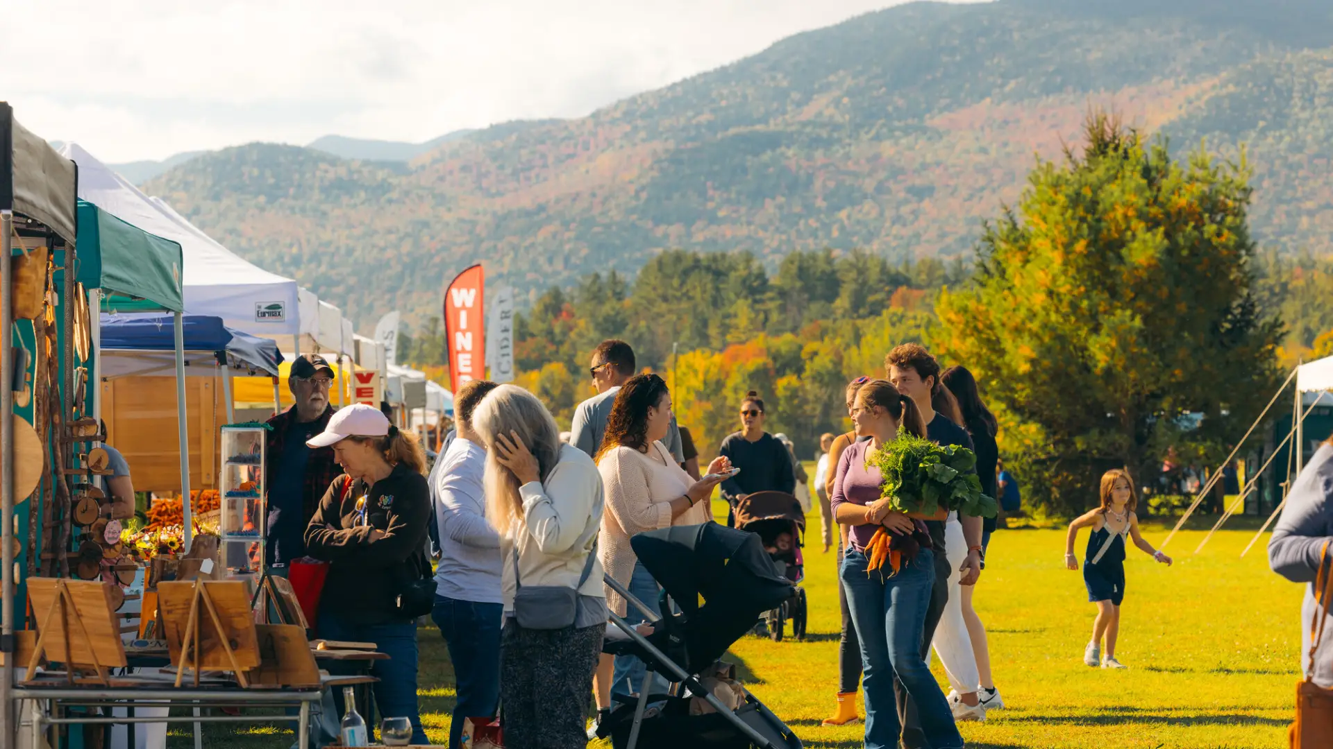 People browse various farmers market vendors and walk between rows of tents with mountains in background
