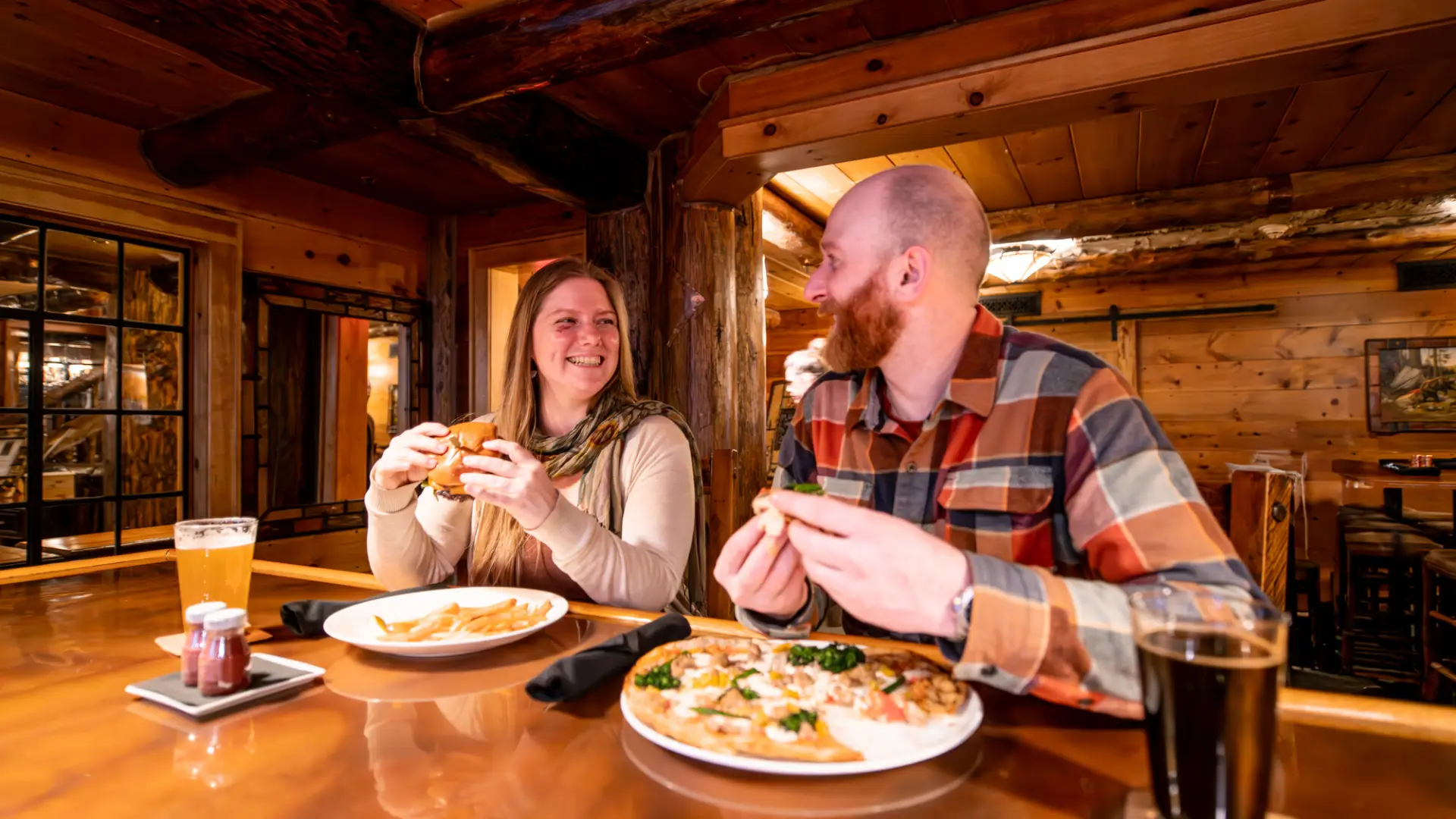 Man and woman sit side-by-side at bar smiling at one another with pints of beer and plates of food holding burgers