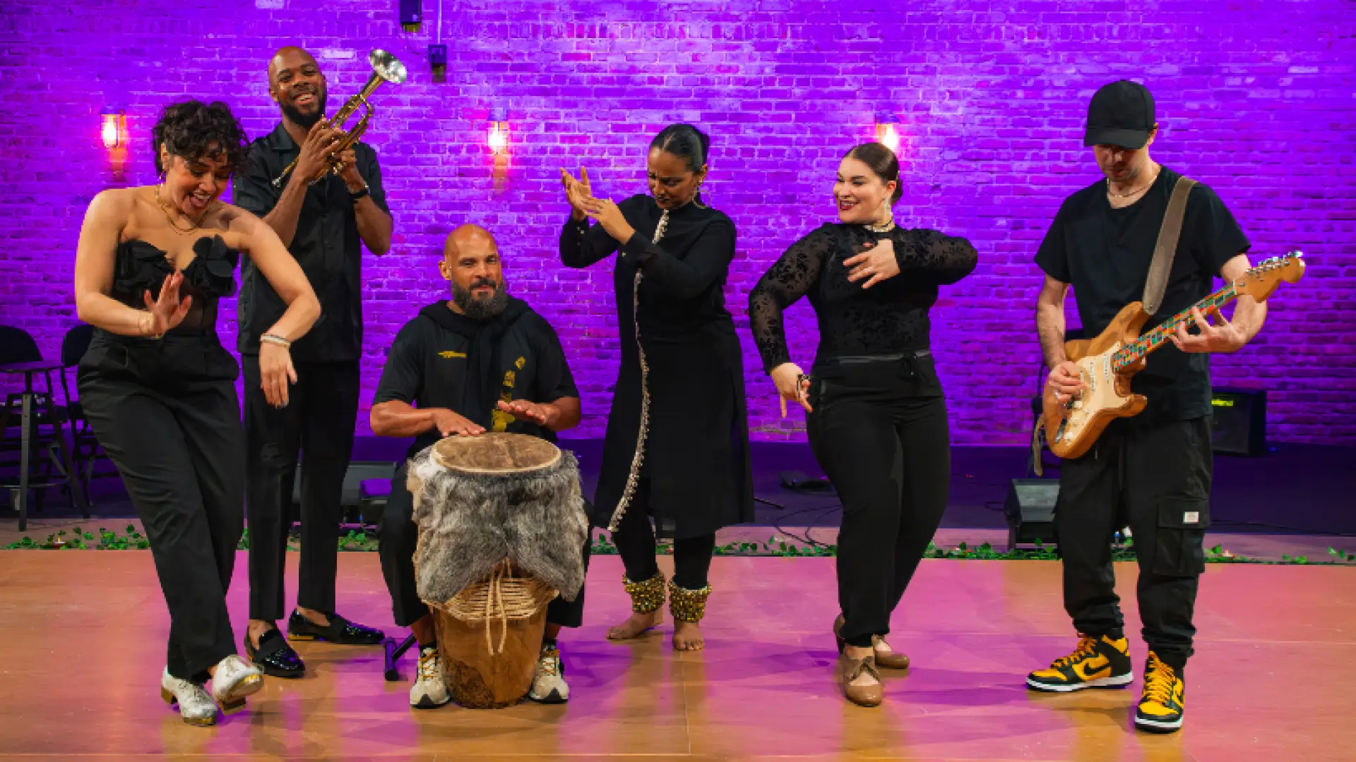 A band comprised of dancers and musicians performs on stage. A woman tap dances in the foreground. Other performers include a trumpet player, a drummer, two female dancers, and a guitarist. Purple lighting illuminates a brick wall in the background.
