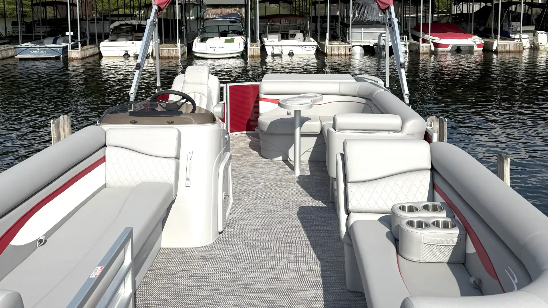 A red and white pontoon boat is docked at the marina with other boats in the covered slips behind