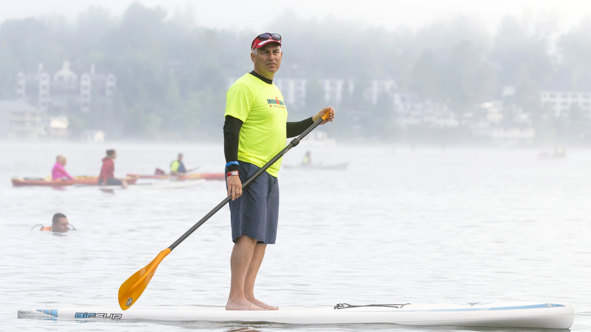 A man in a bright yellow Volunteer tshirt and shorts stands on a paddle board on a misty morning watching a race