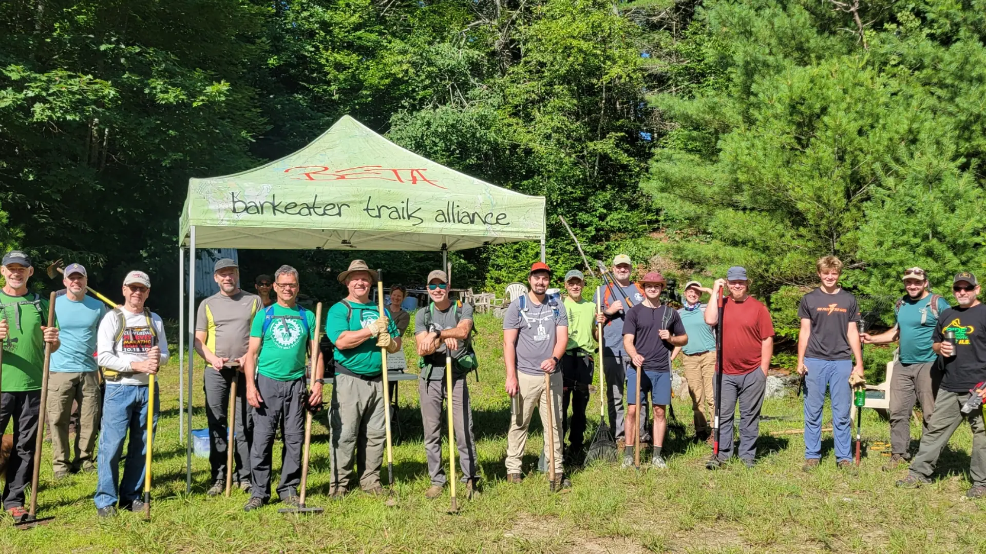 A group of volunteers stand in front of a Barkeater Trails Alliance tent, holding trail maintenance tools. 
