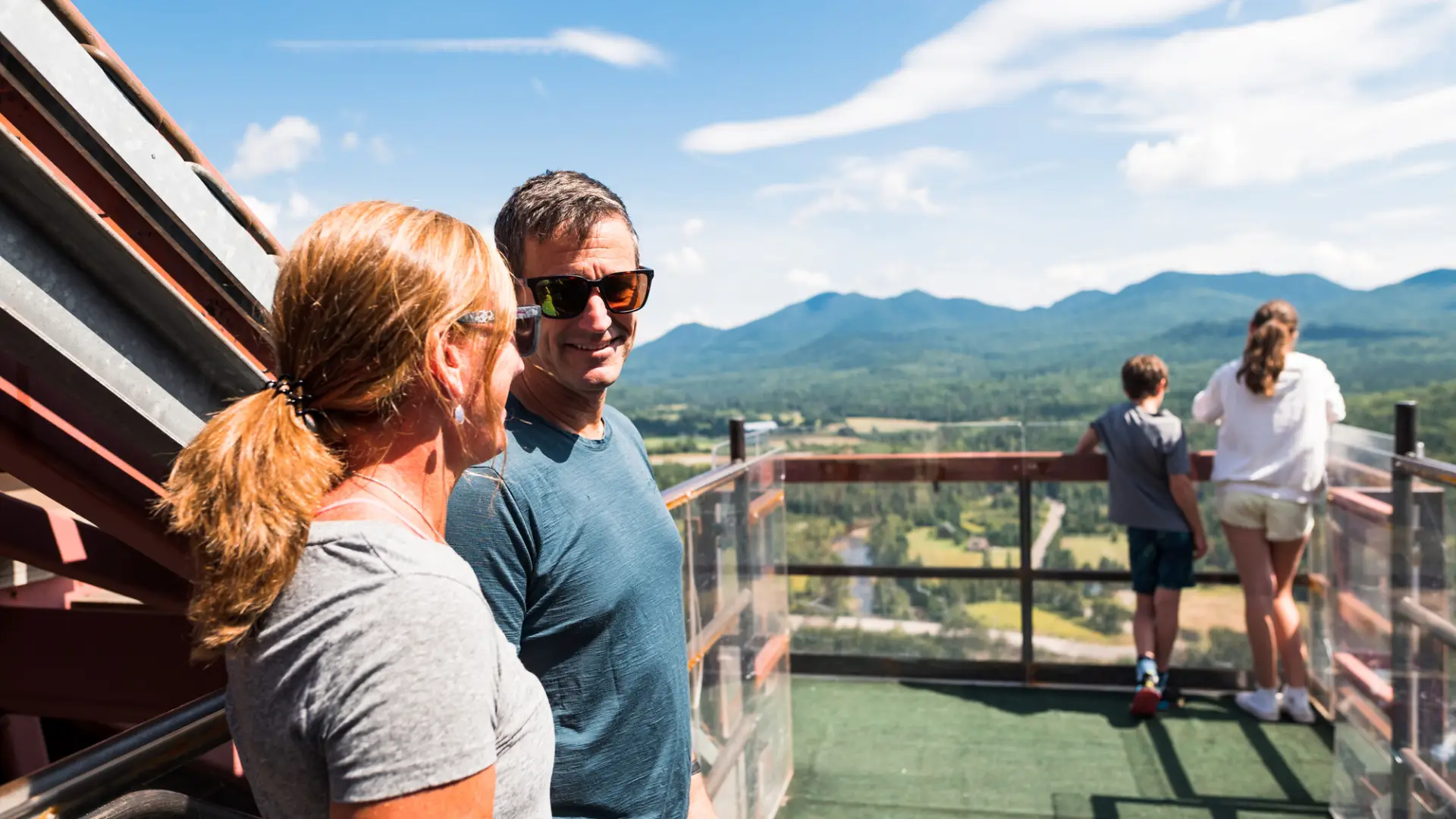 A man and woman smile to each other while their children overlook Olympic ski jumps.