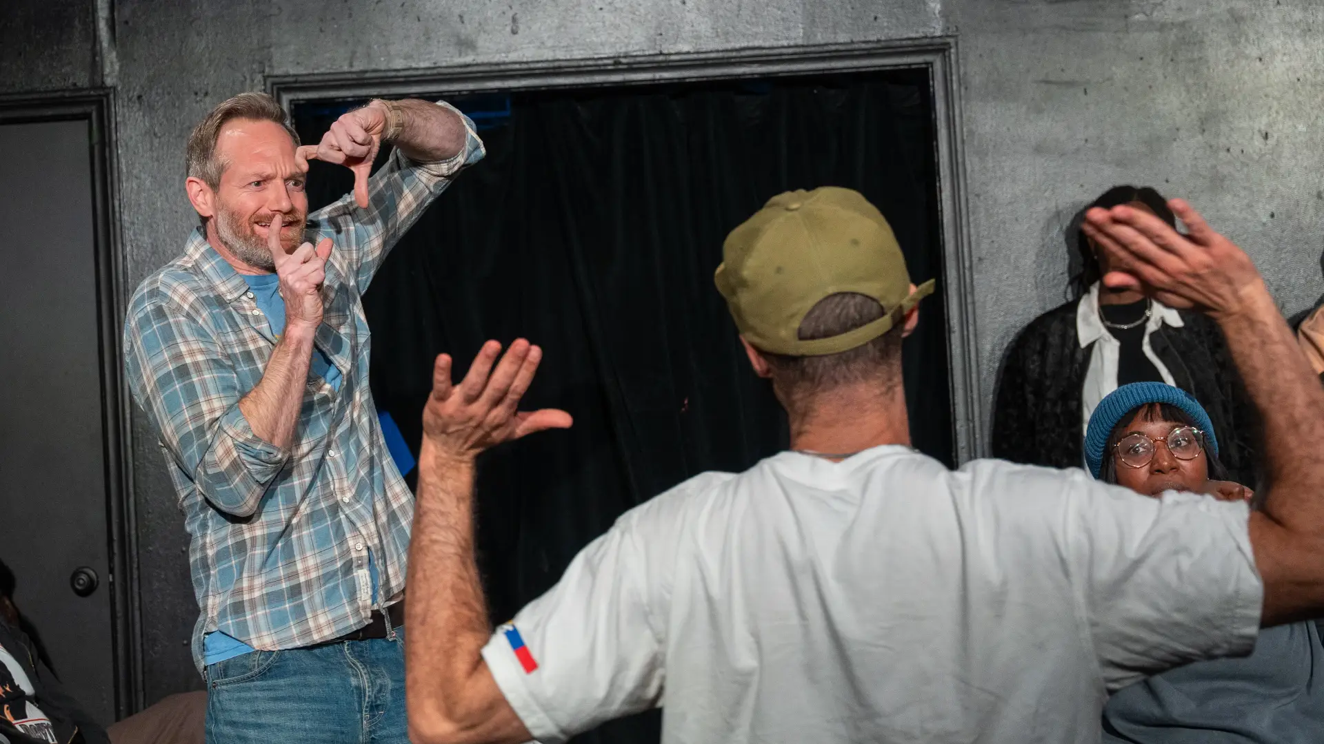 Male improv actor makes box with fingers and looks skeptically through it at person with back to frame holding up arms