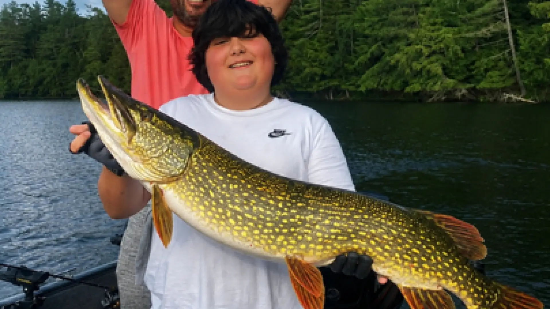A kid holds a large pike while a man throws up his hands in victory behind.